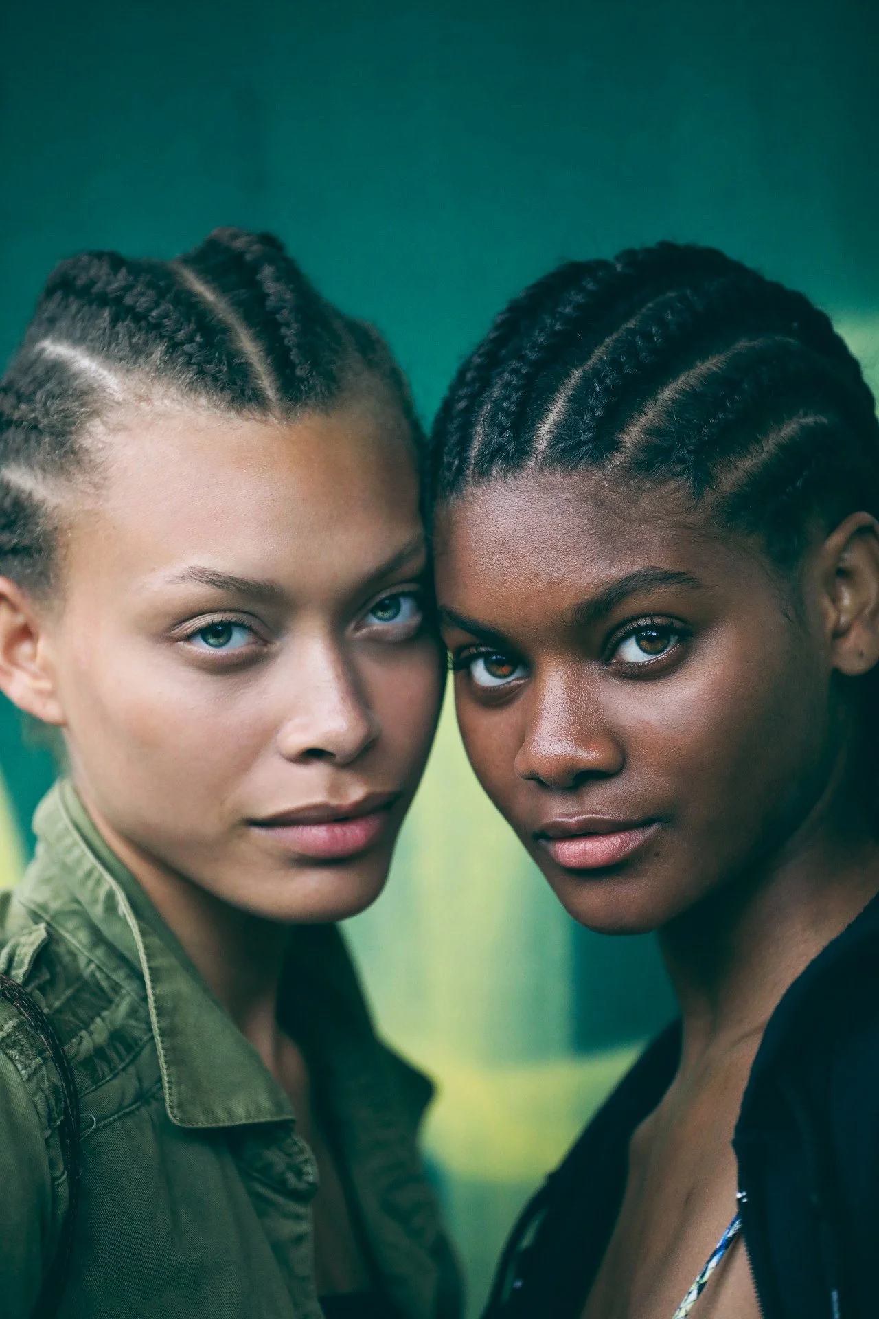 Two women with braided hairstyles and serious expressions standing close together with a blurred green background.