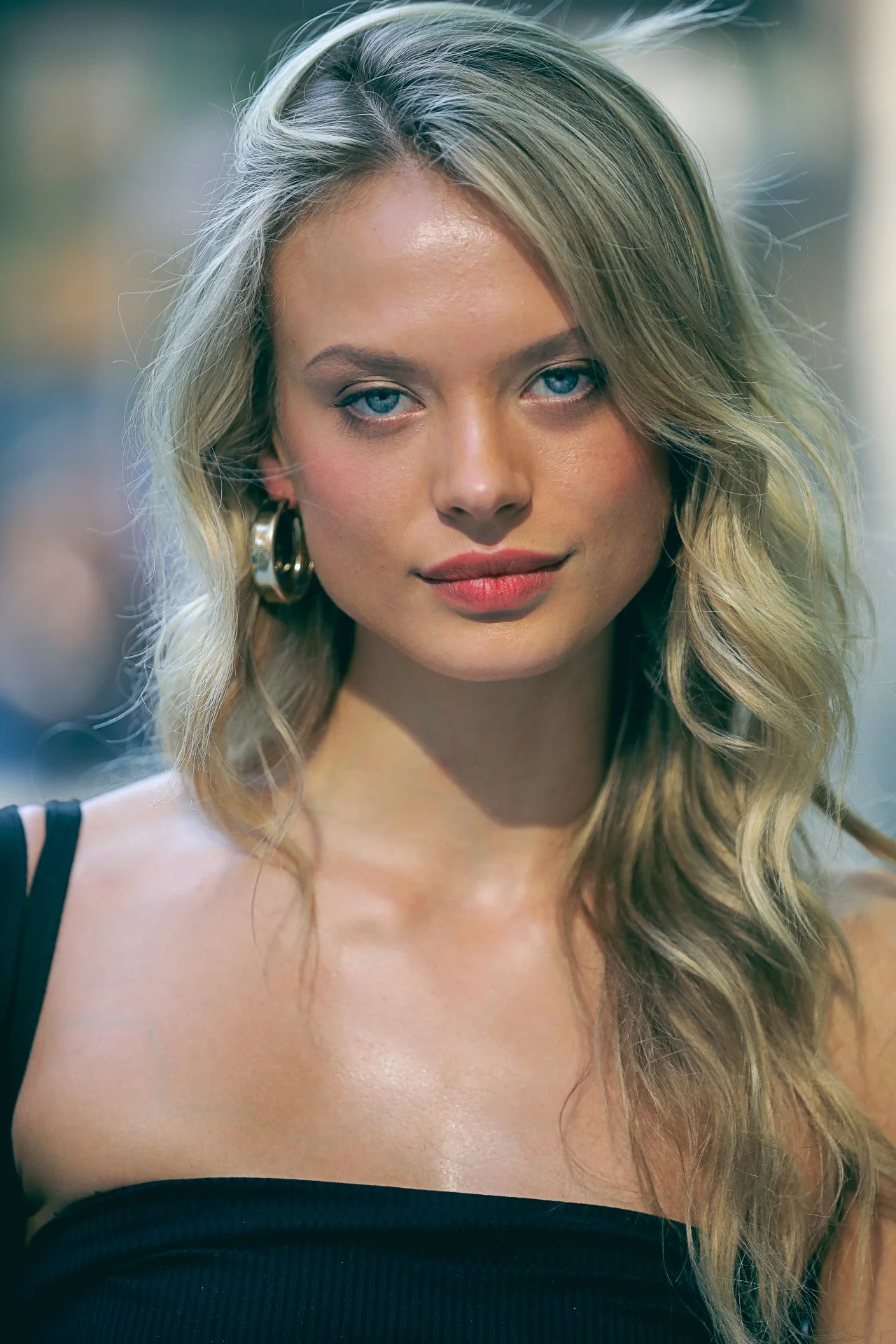 A young woman with blonde, wavy hair, blue eyes, and hoop earrings, wearing a black sleeveless top, smiling softly outdoors.
