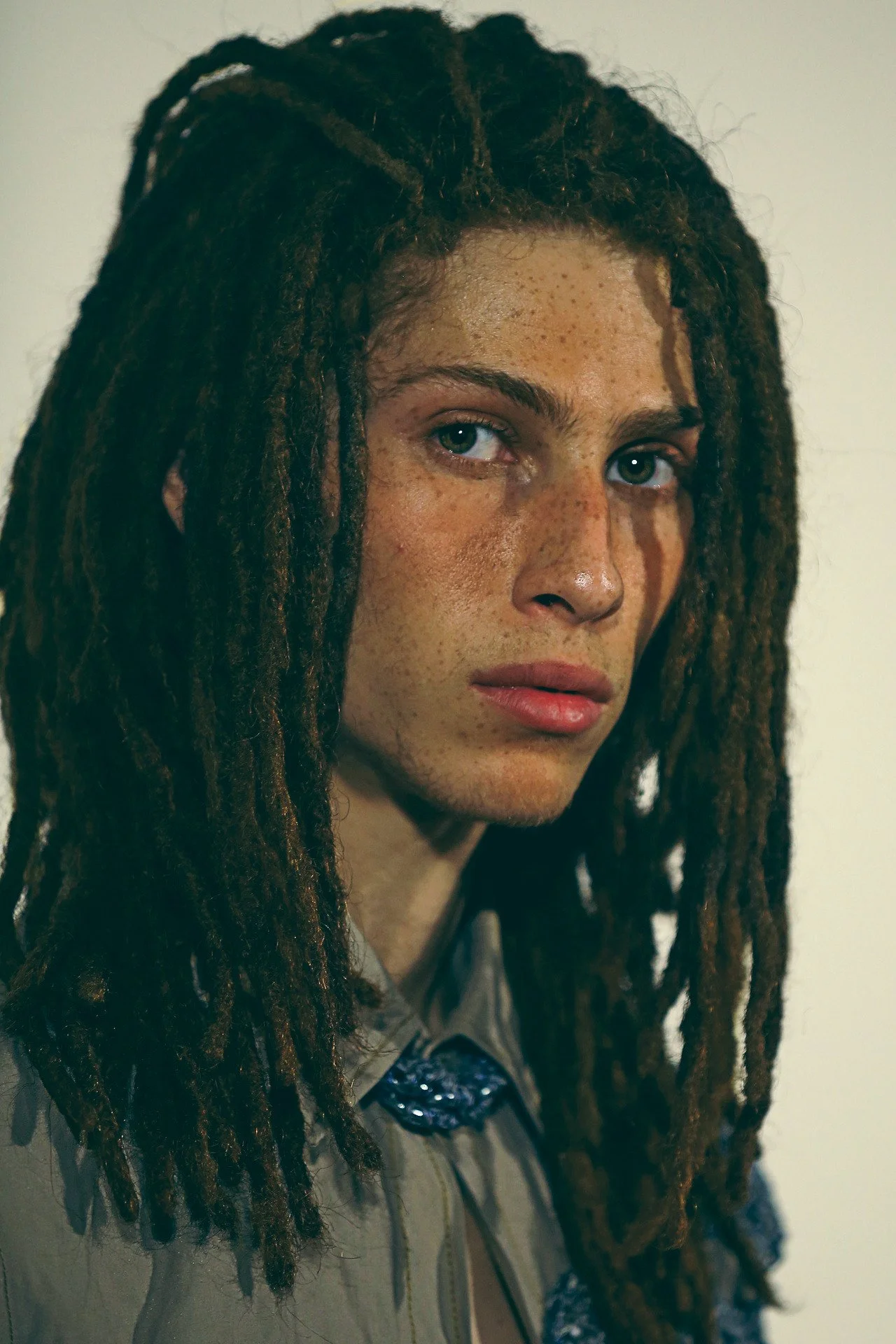 A woman with long dreadlocks, light skin, and freckles faces the camera against a neutral background.