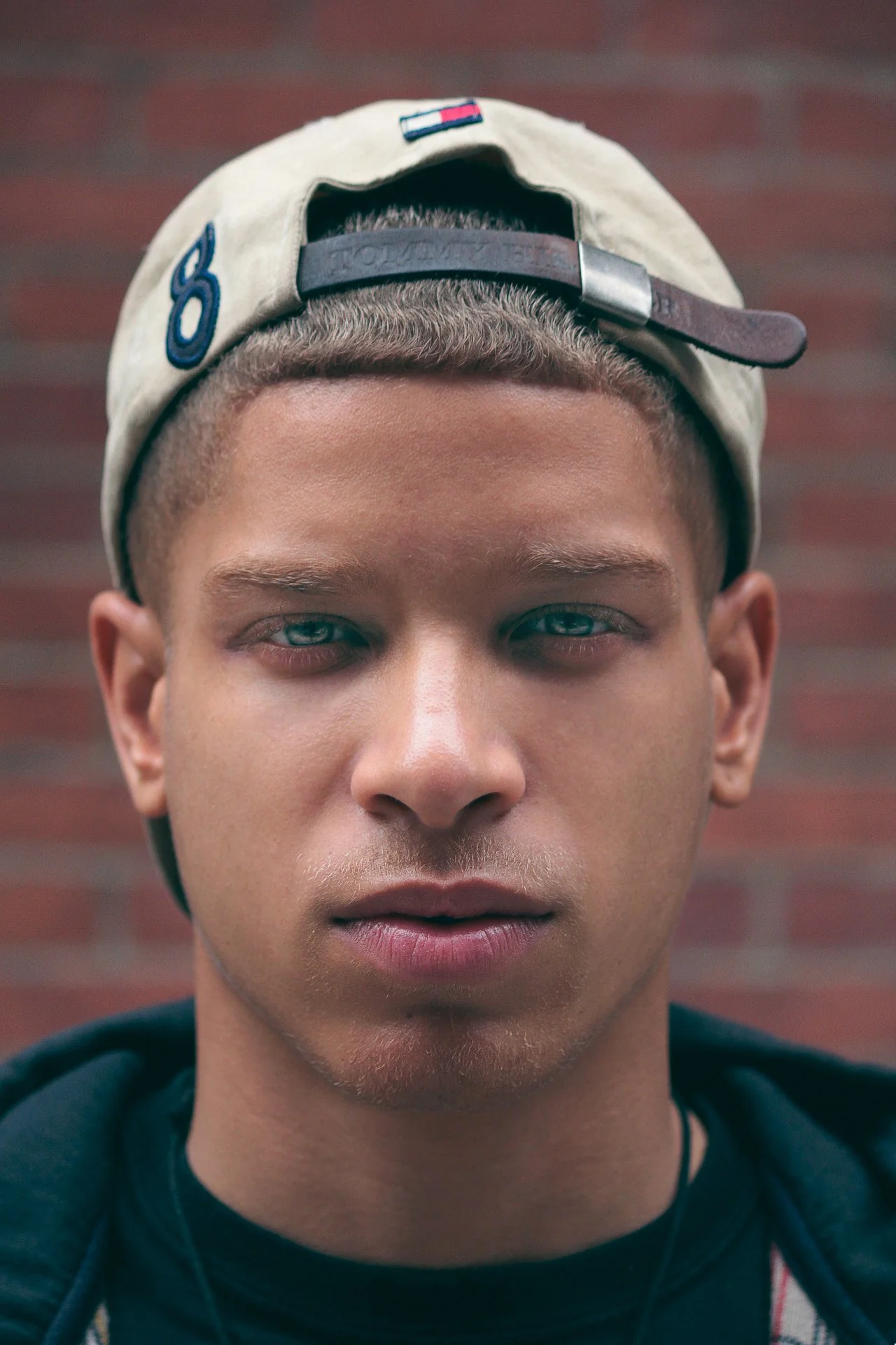 Close-up of a young man wearing a beige baseball cap backwards, with a brick wall in the background.