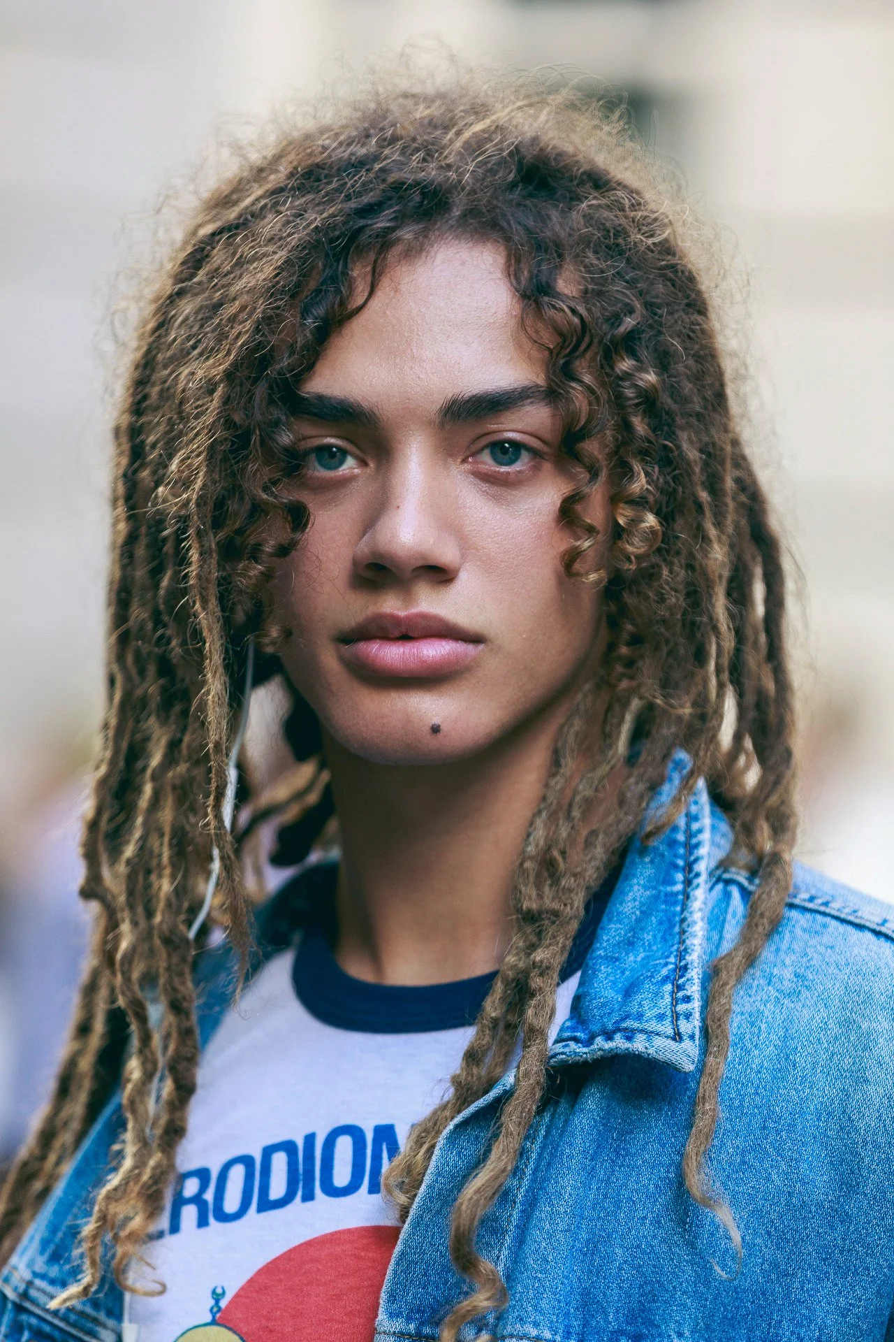 Close-up of a young woman with light skin, blue eyes, and long messy dreadlocks, wearing a denim jacket over a white T-shirt with colorful graphic design.
