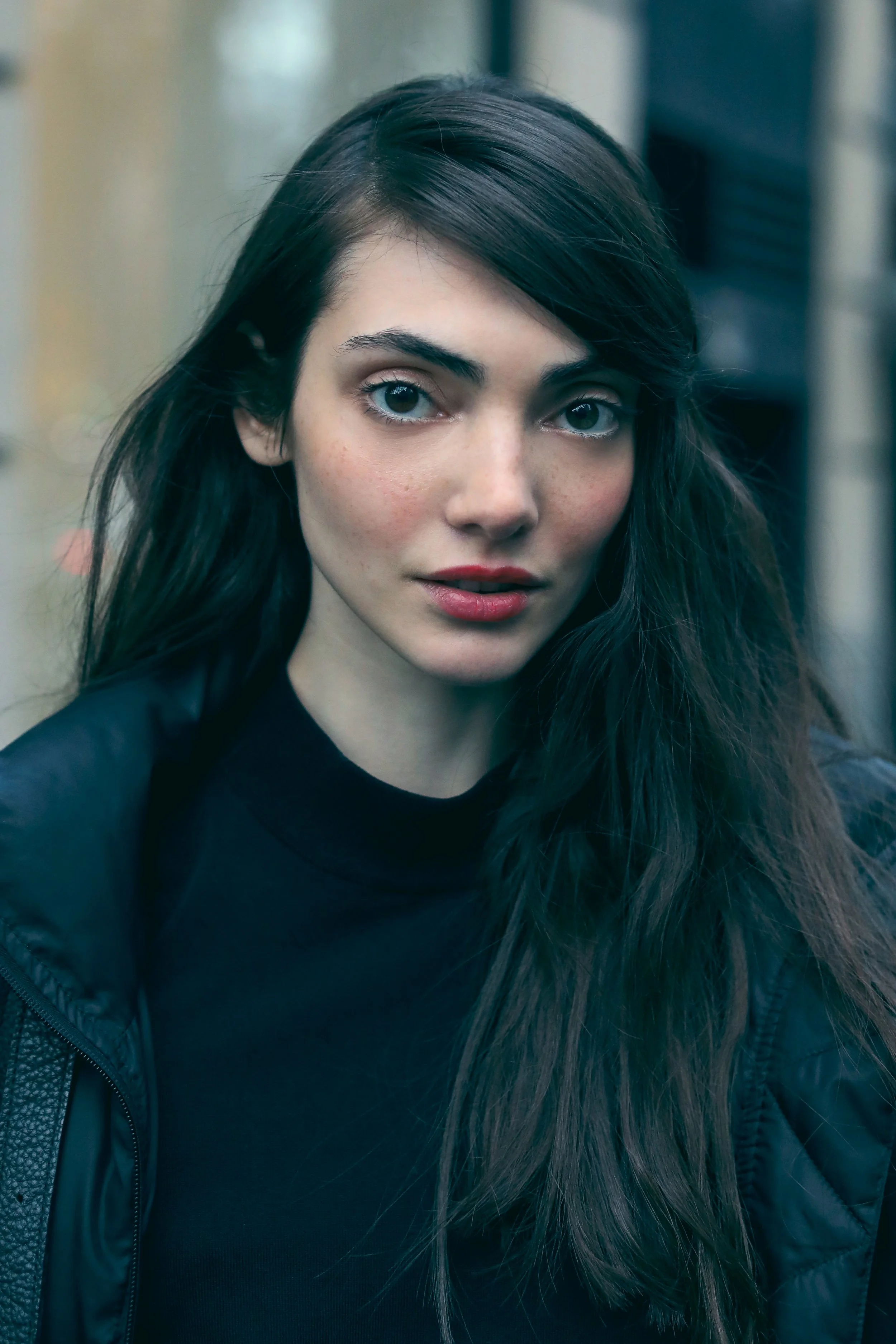 Young woman with long dark hair, fair skin, and blue eyes, wearing a black top and black jacket, standing outdoors with blurred urban background.