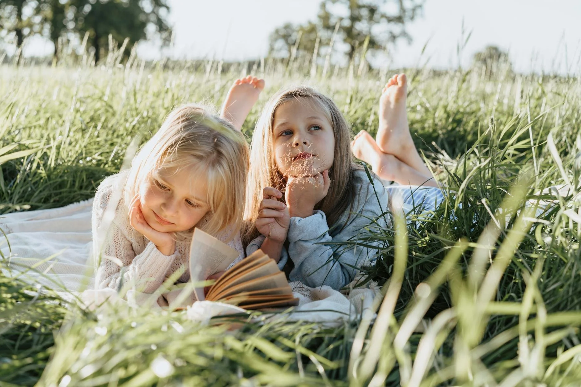 Two young girls lying on a blanket in a sunlit grassy field, looking at a book together while enjoying a summer afternoon outdoors.
