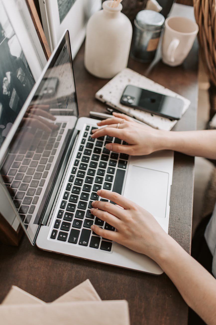 A top-down view of a person's hands typing on a laptop keyboard at a wooden desk with a smartphone nearby.