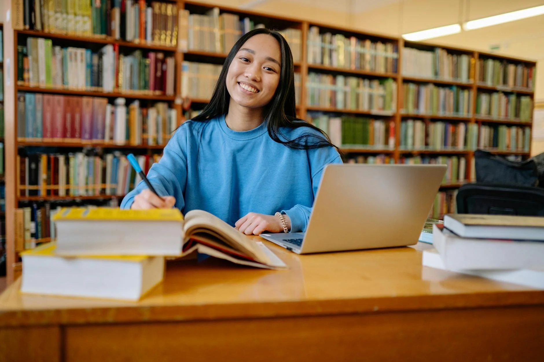A smiling young woman sitting at a desk in a library with a laptop and open books, with bookshelves in the background.