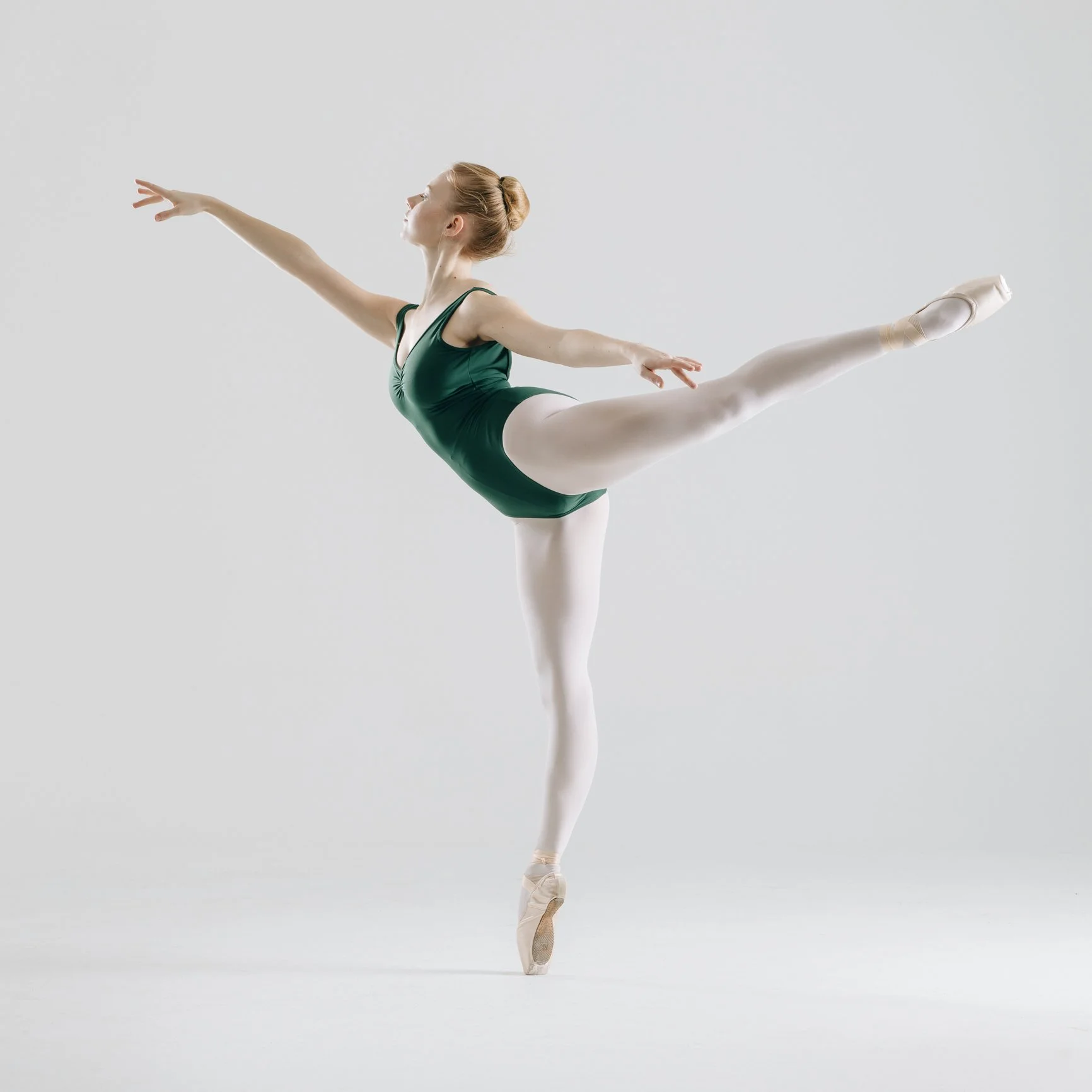 Ballet dancer in a green leotard on pointe, balancing on one foot with other leg extended behind her, arms outstretched.