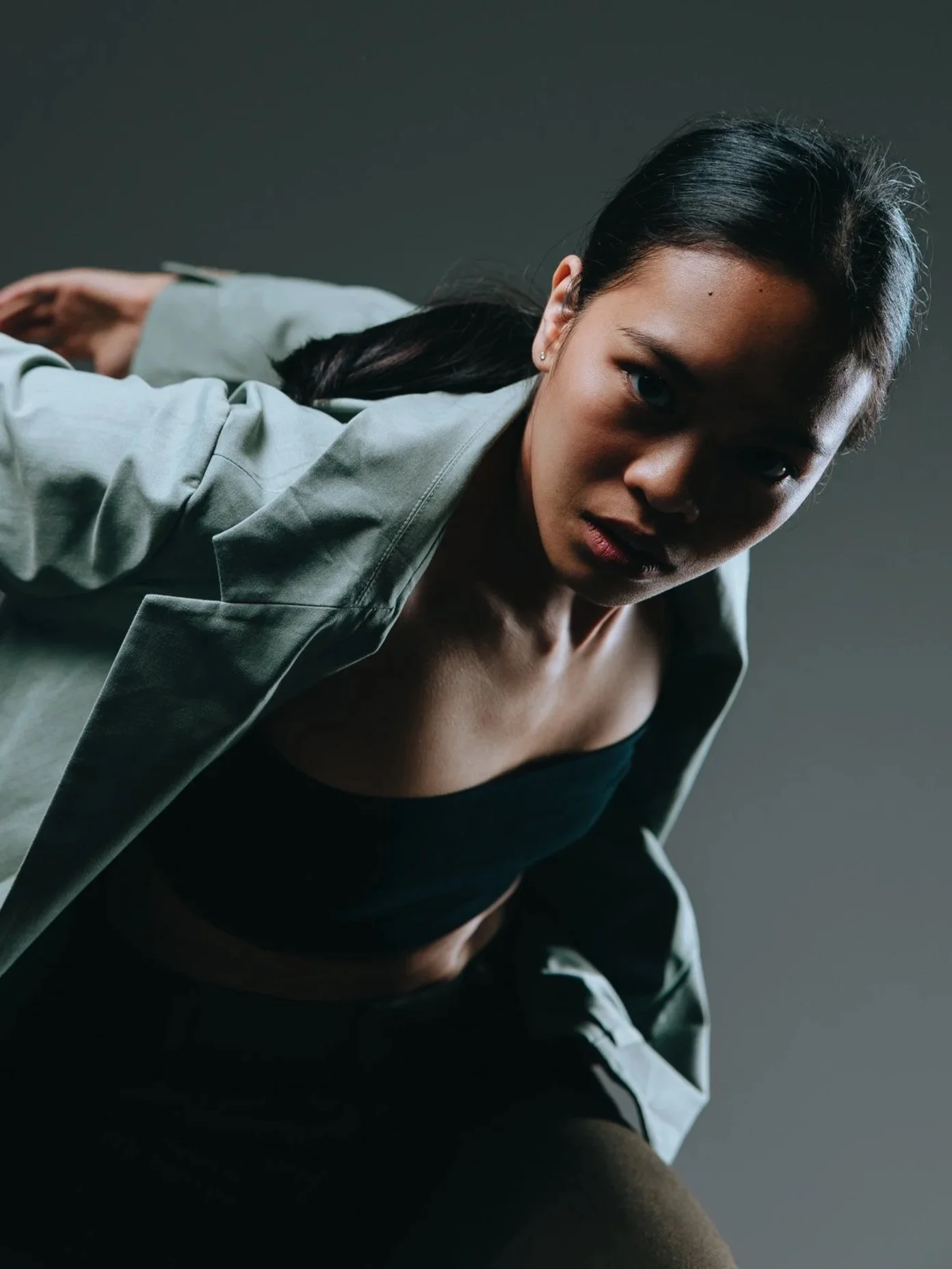 A woman with dark hair pulled back, wearing a light-colored blazer over a black crop top, leans forward with a serious expression against a dark background.