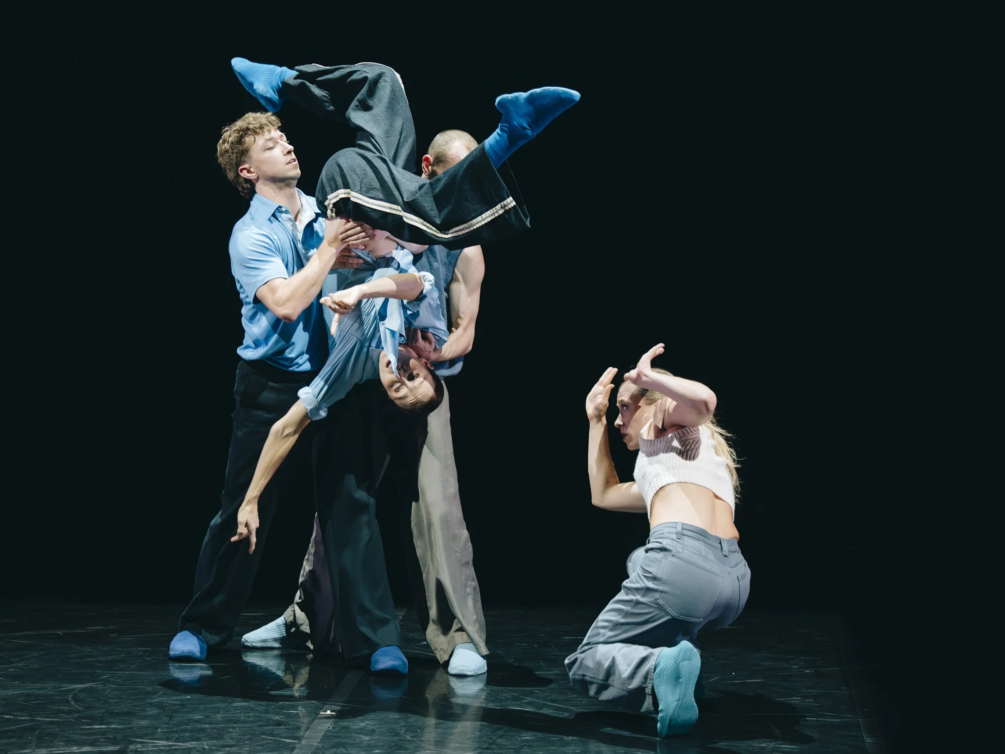 A group of five dancers performing on stage, with one dancer upside down and others supporting and engaging with her, against a black background.