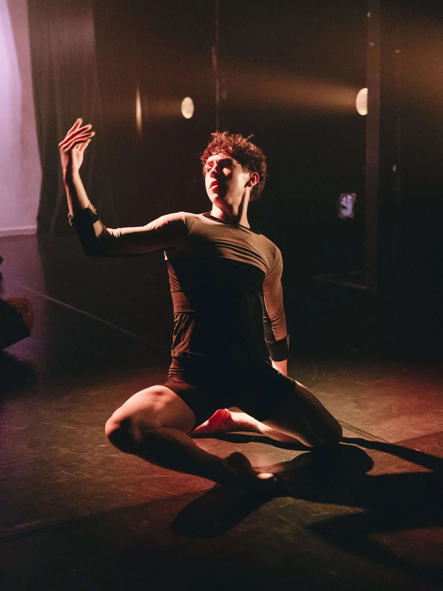 A person performing a dance or dramatic pose on stage, kneeling with one leg bent and the other extended, with one arm raised and the other resting on their knee, illuminated by stage lighting.