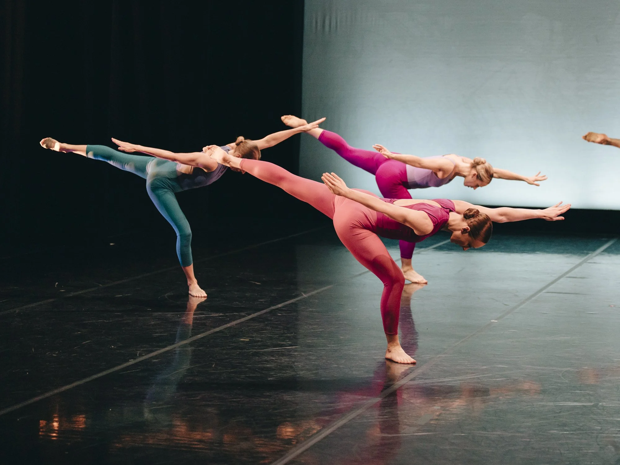 Four women in colorful workout clothes practicing ballet or yoga on stage, balancing on one leg with arms extended and other leg raised behind them.