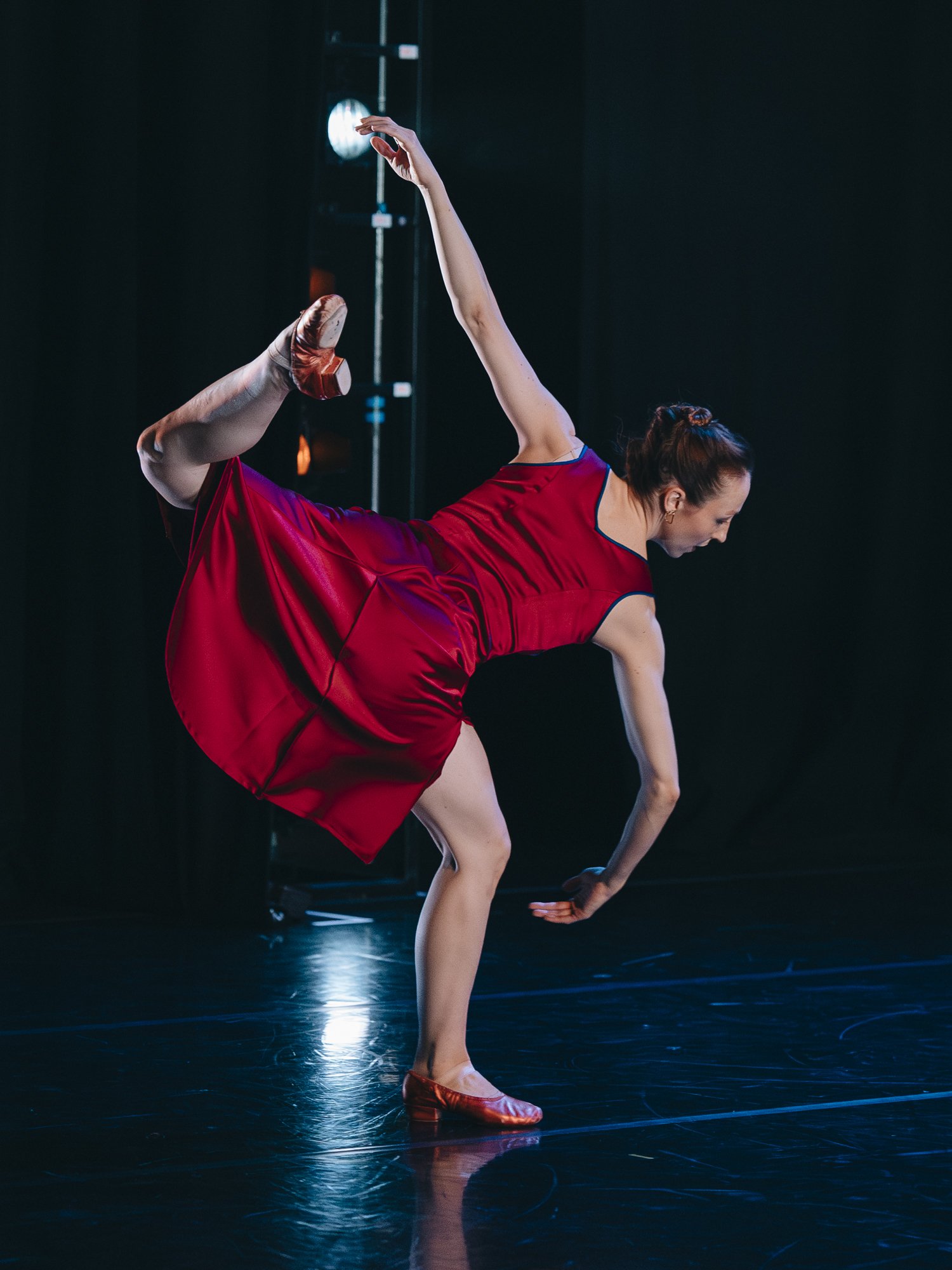 A female dancer performing a contemporary dance move on stage, wearing a red dress and ballet shoes, with one leg raised behind her and her arms extended in a graceful pose.