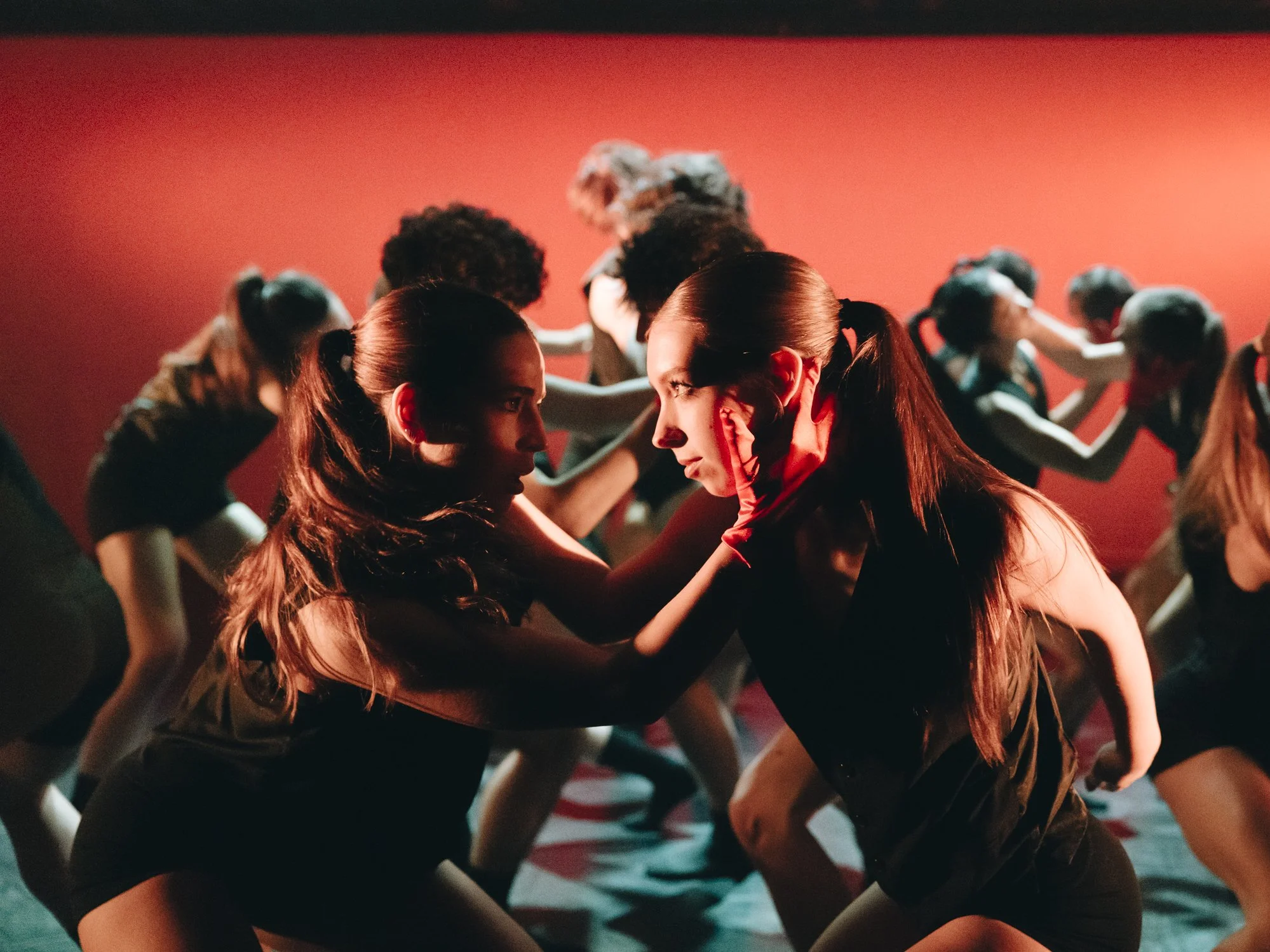 Two women with ponytails face each other in a dance or theatrical performance, with a group of dancers behind them, all in black outfits and striking dramatic poses against a red background.