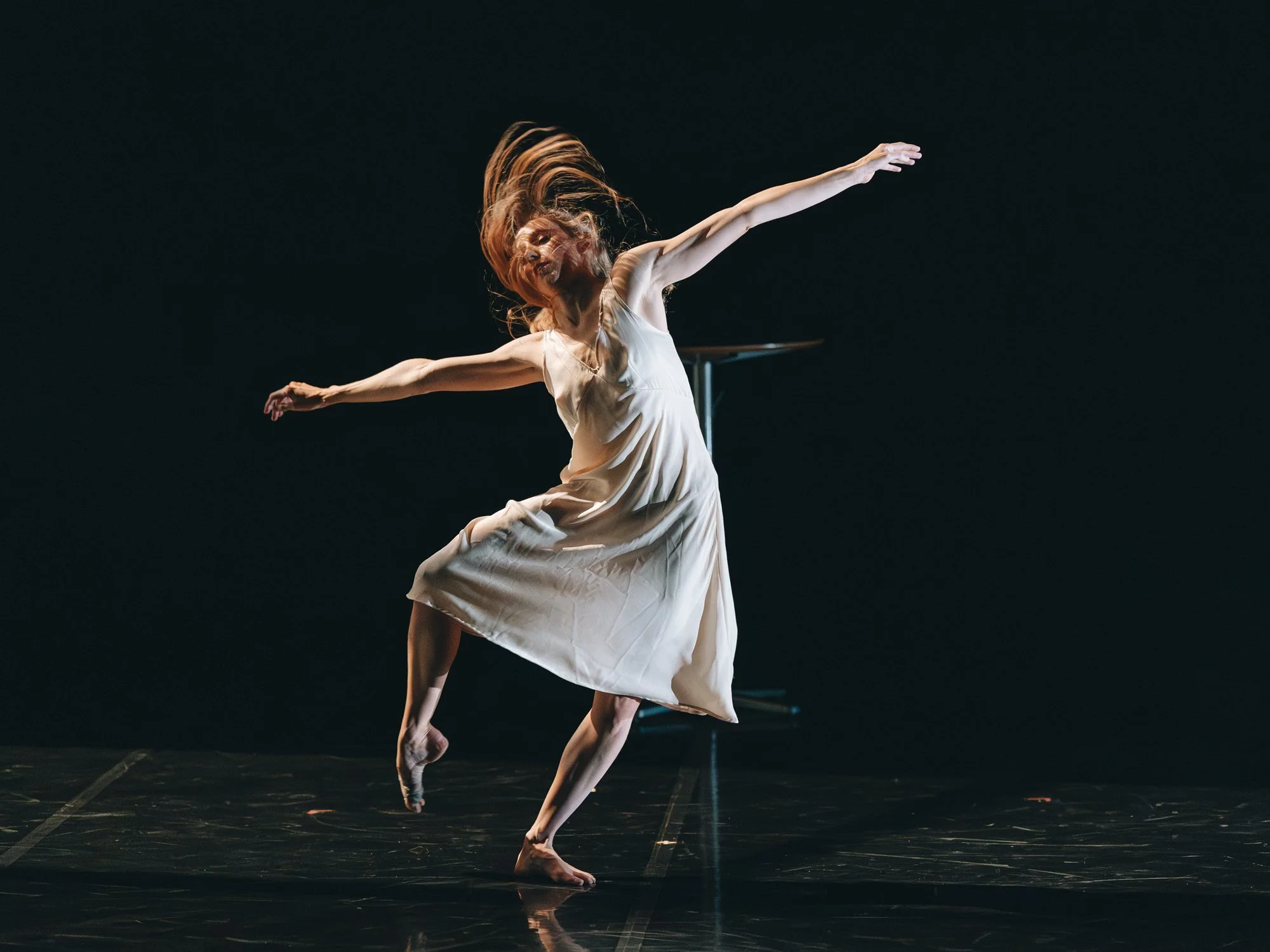 A female ballet dancer in a white dress performing a dance move on stage with a dark background.
