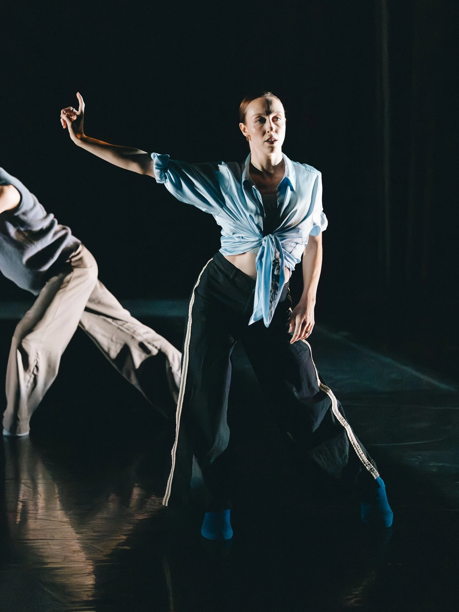 Dancer performing on stage wearing a light blue tied shirt and black pants with white stripes, with another dancer partially visible beside them.