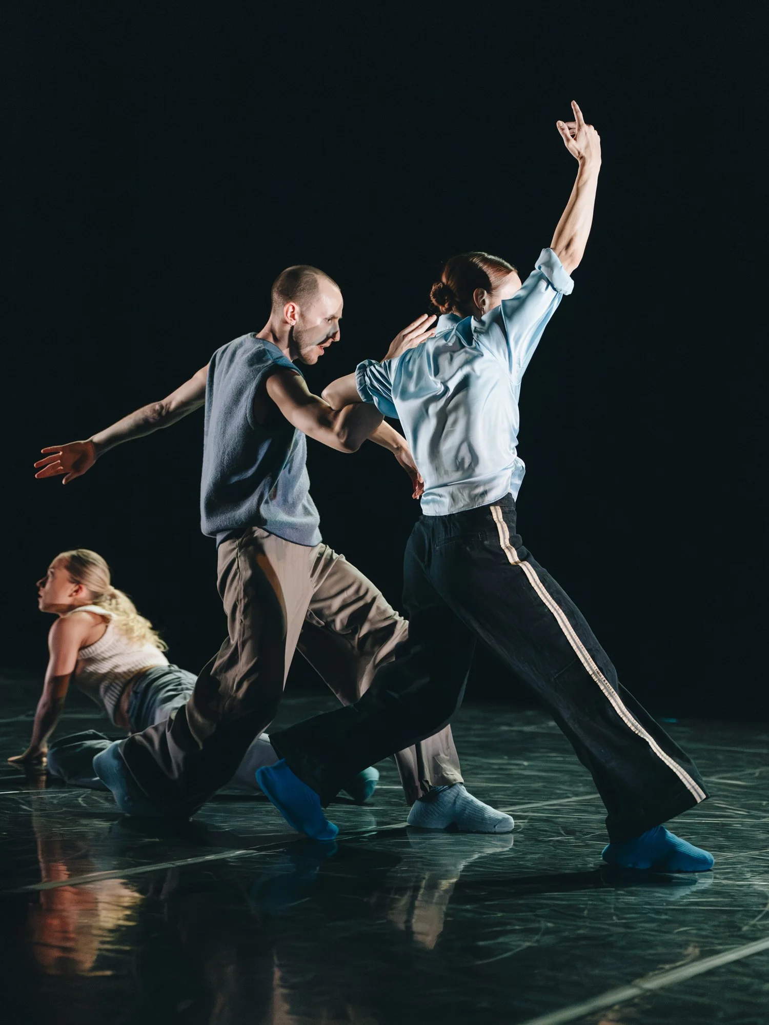 Three dancers perform on a dark stage, with two standing and one kneeling on the floor, capturing an expressive dance pose.
