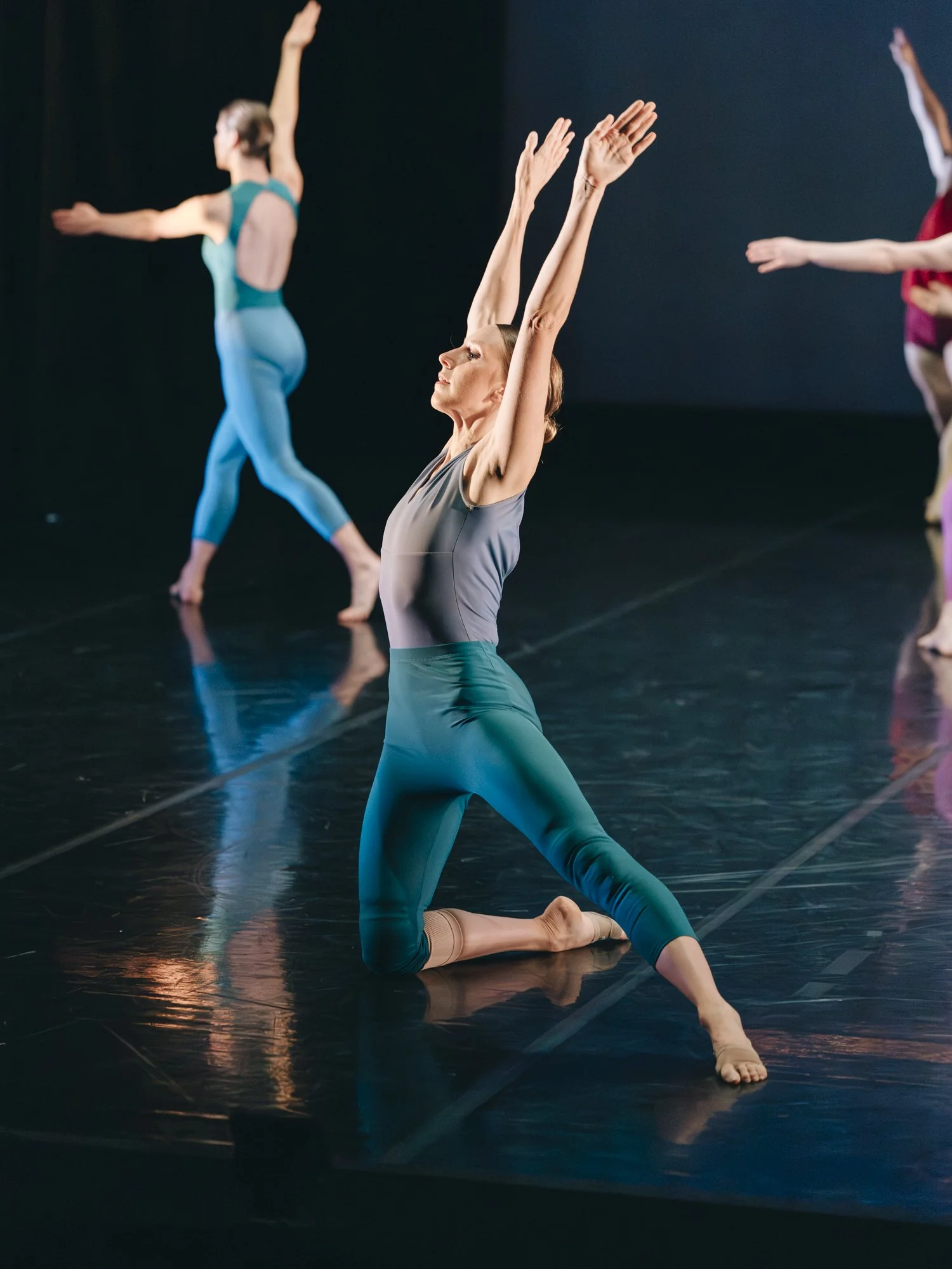 A ballet dancer in a gray top and teal leggings kneels on stage with arms raised overhead, other dancers in colorful costumes perform in the background.