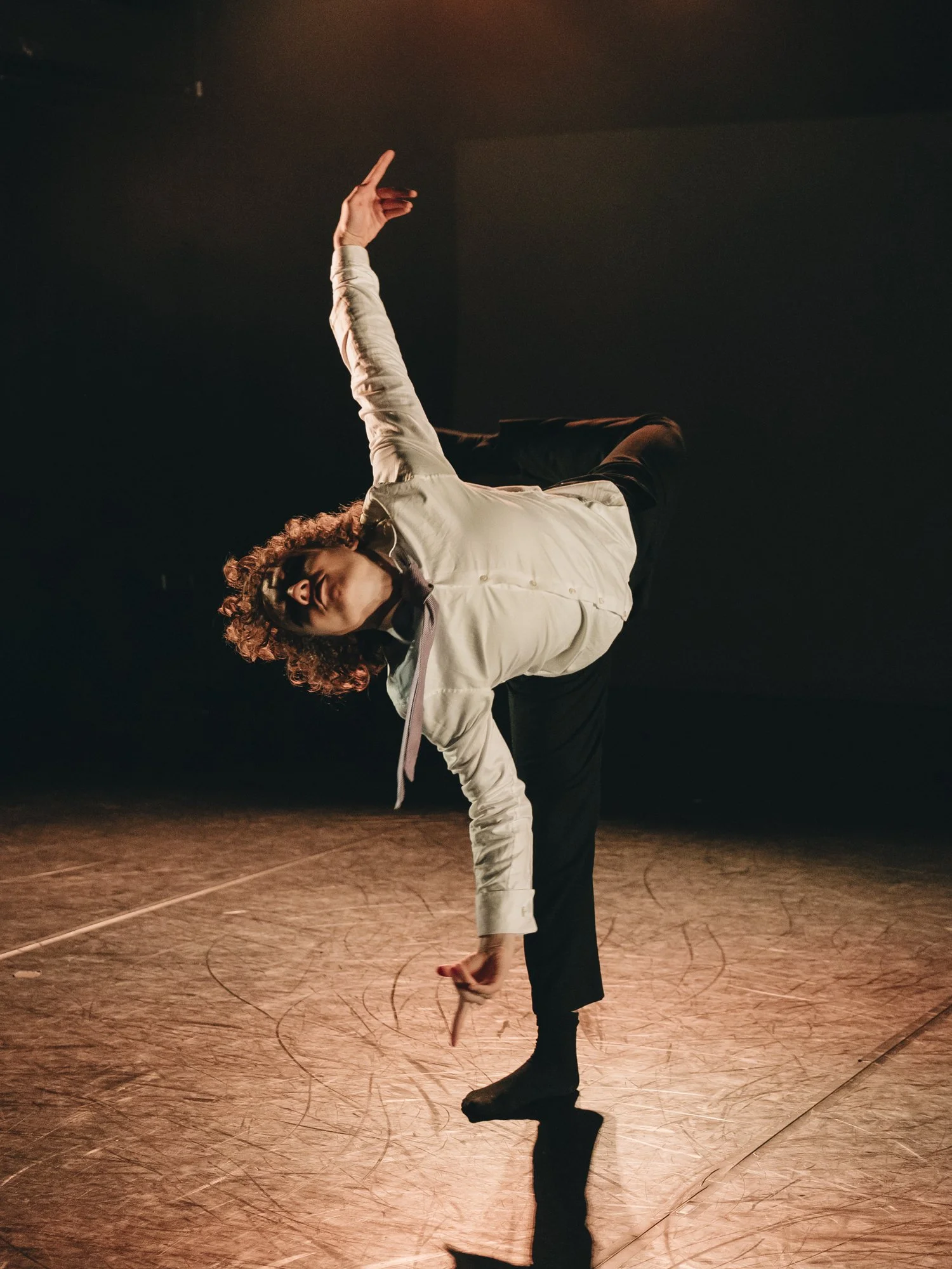 A dancer performs a balancing pose on a dimly lit stage, leaning forward with one leg extended and arms raised in a graceful arc.