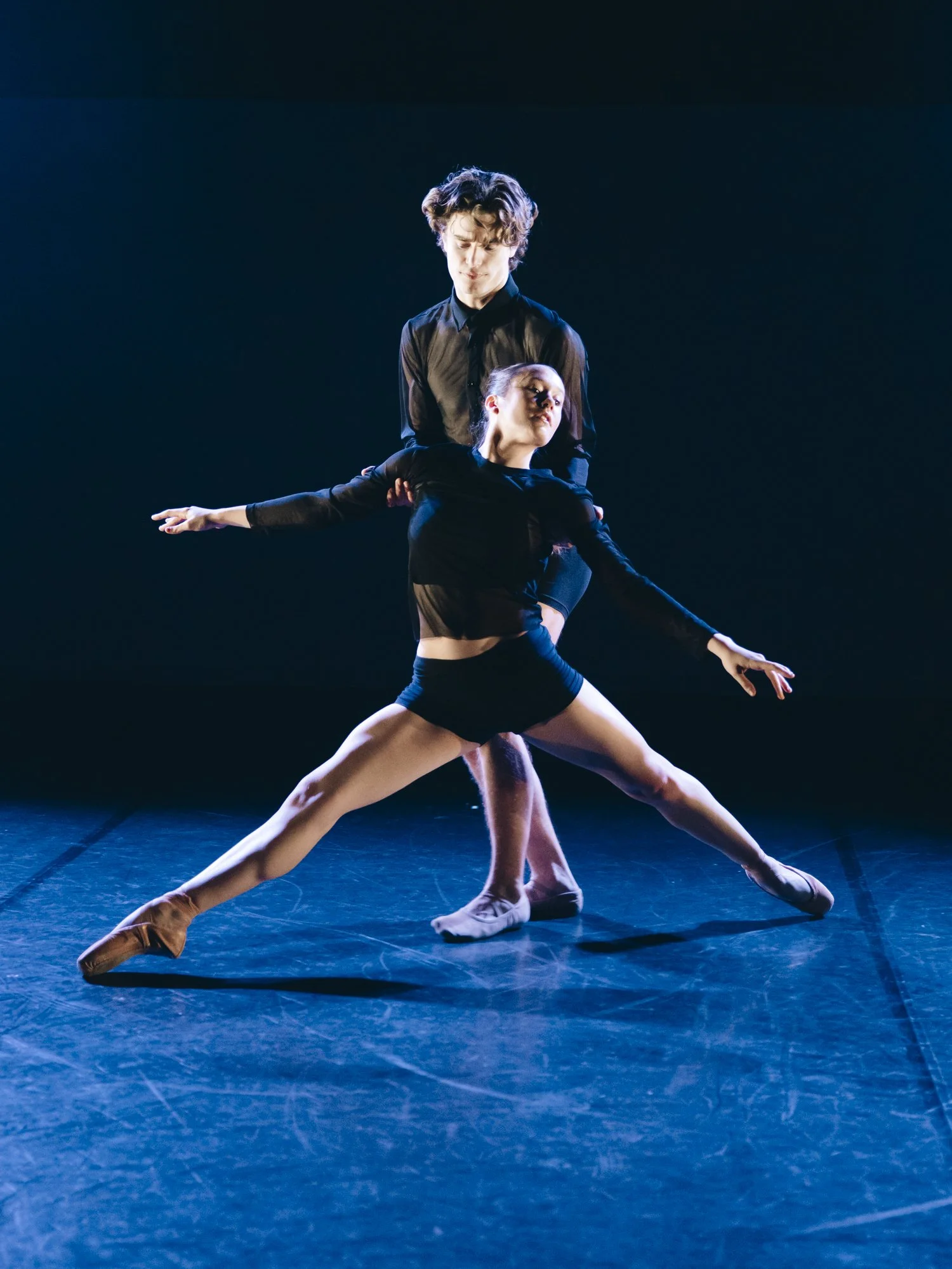Two ballet dancers performing a duet on stage, with one supporting the other. The female dancer is in a deep side split with arms extended, and the male dancer stands behind her, holding her waist. Both are dressed in black and are illuminated by sta