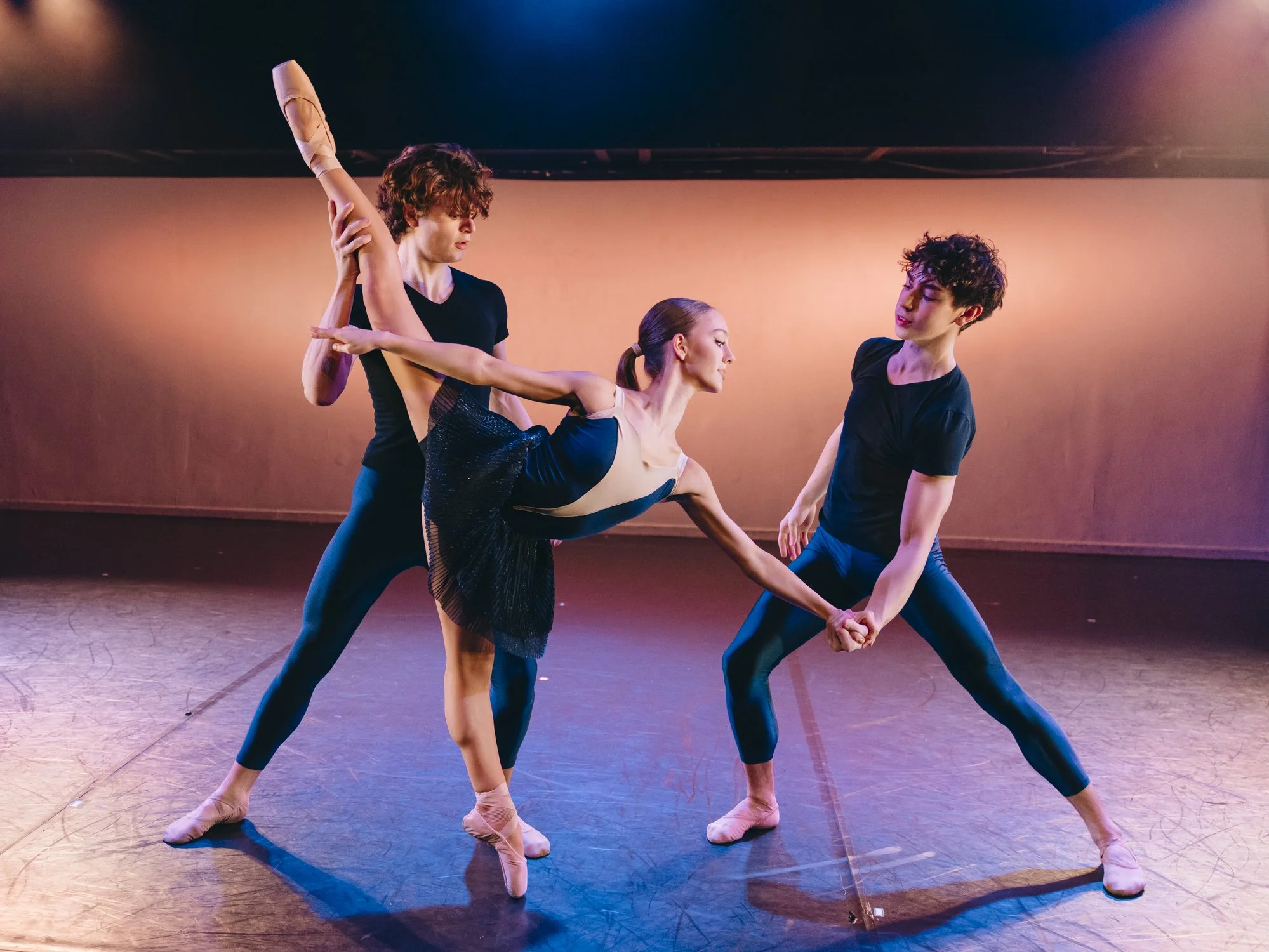 Three ballet dancers practicing on stage, two men and one woman, with the woman being lifted in a dance pose supported by the two men.