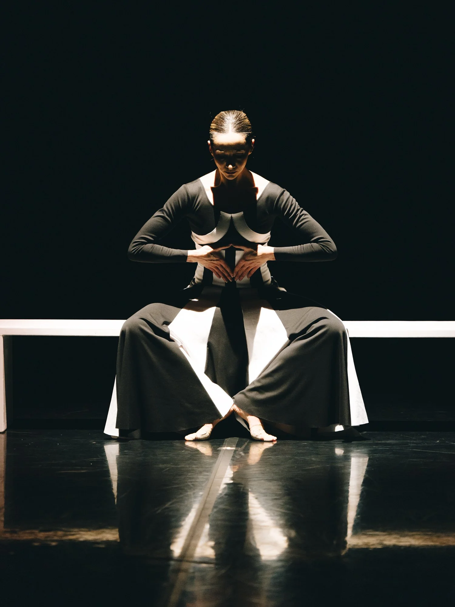 A woman in black and white clothing sits on a bench with her arms crossed in front of her, against a dark background, with her reflection visible on the shiny floor.