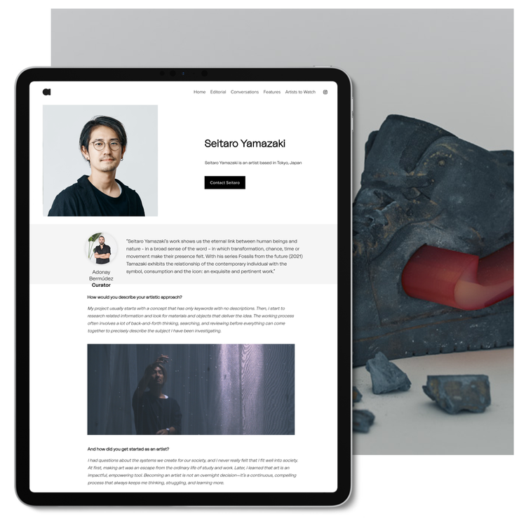 A tablet displaying an artist's online profile with a portrait, biography, and interview, placed on a desk next to a sculpture and rocks.