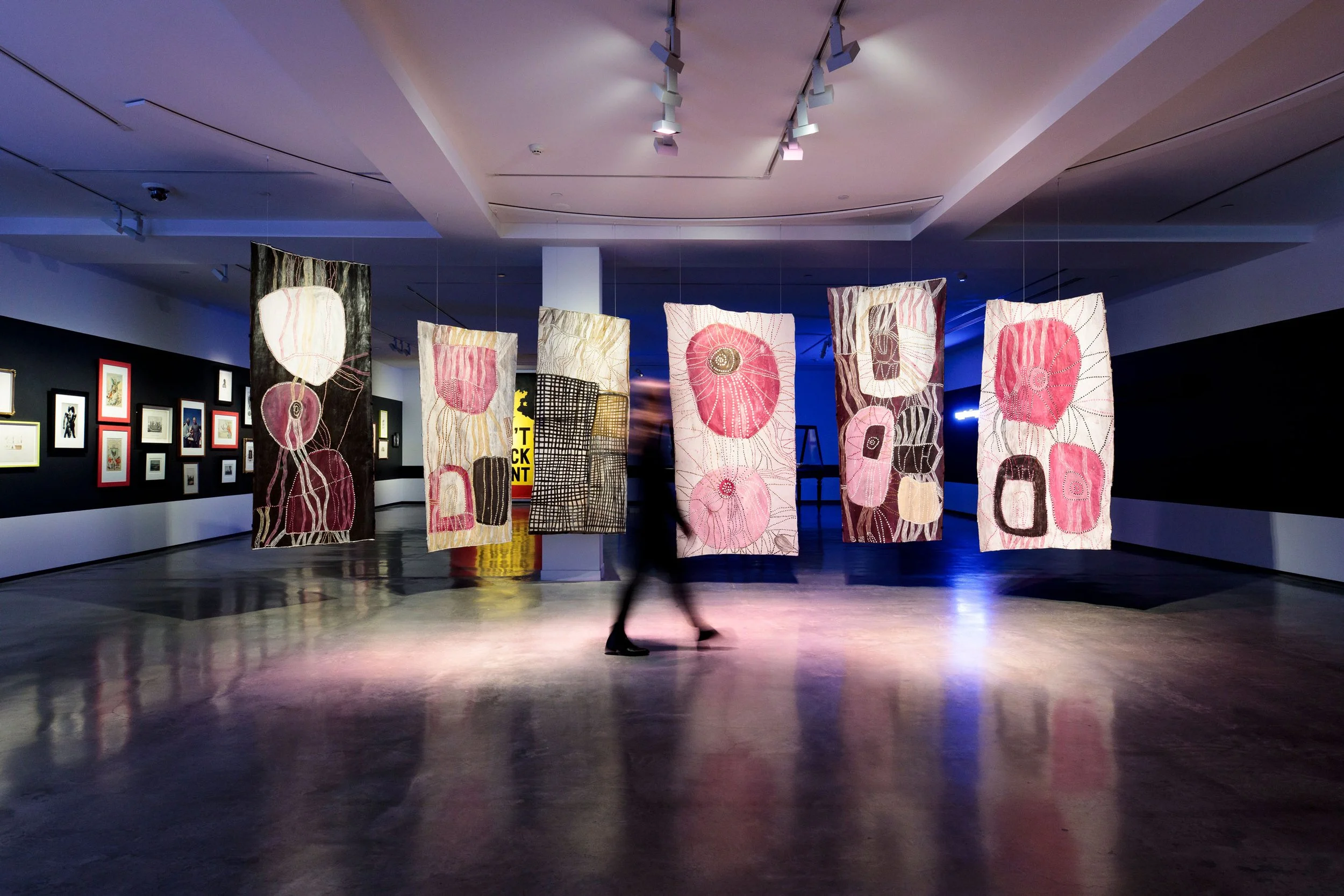 Art gallery interior with hanging fabric tapestries featuring abstract floral and geometric designs in pink, black, and white, with framed artwork on dark walls, and a person walking through the space.