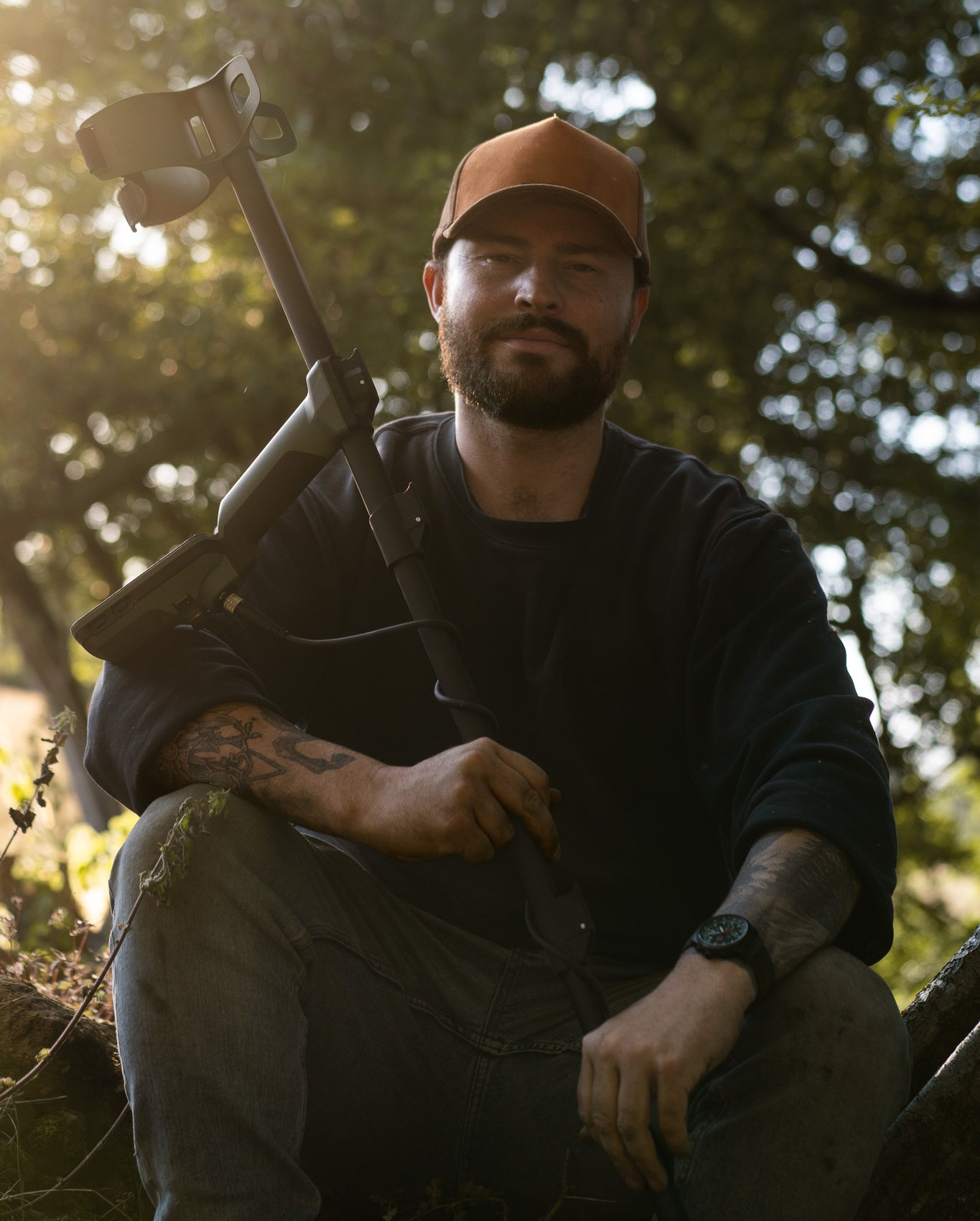 A man sitting outdoors in a wooded area, holding a metal detector, wearing a brown cap, blue sweatshirt, jeans, and a watch, with sunlight filtering through the trees in the background.
