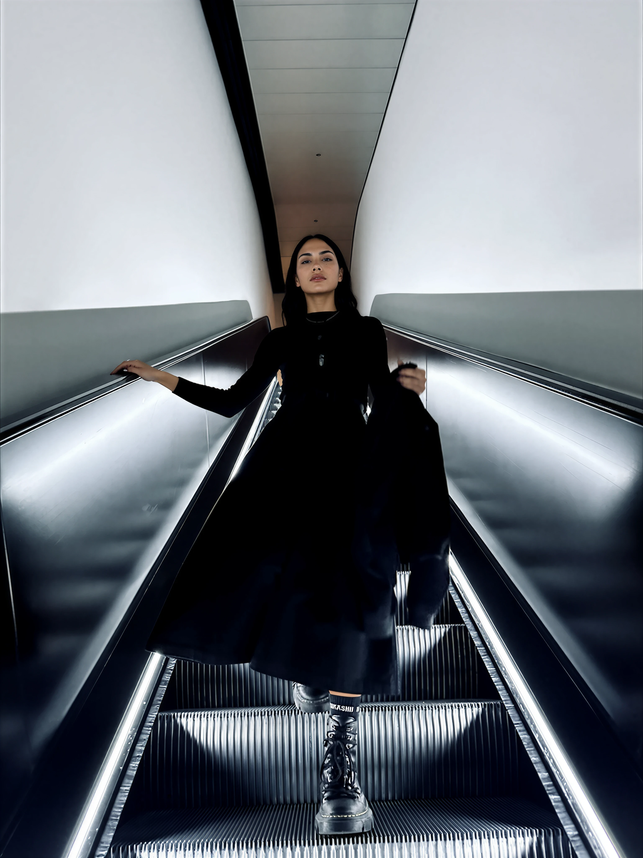 A woman in black clothing and black boots riding an escalator in a modern, sleek building.