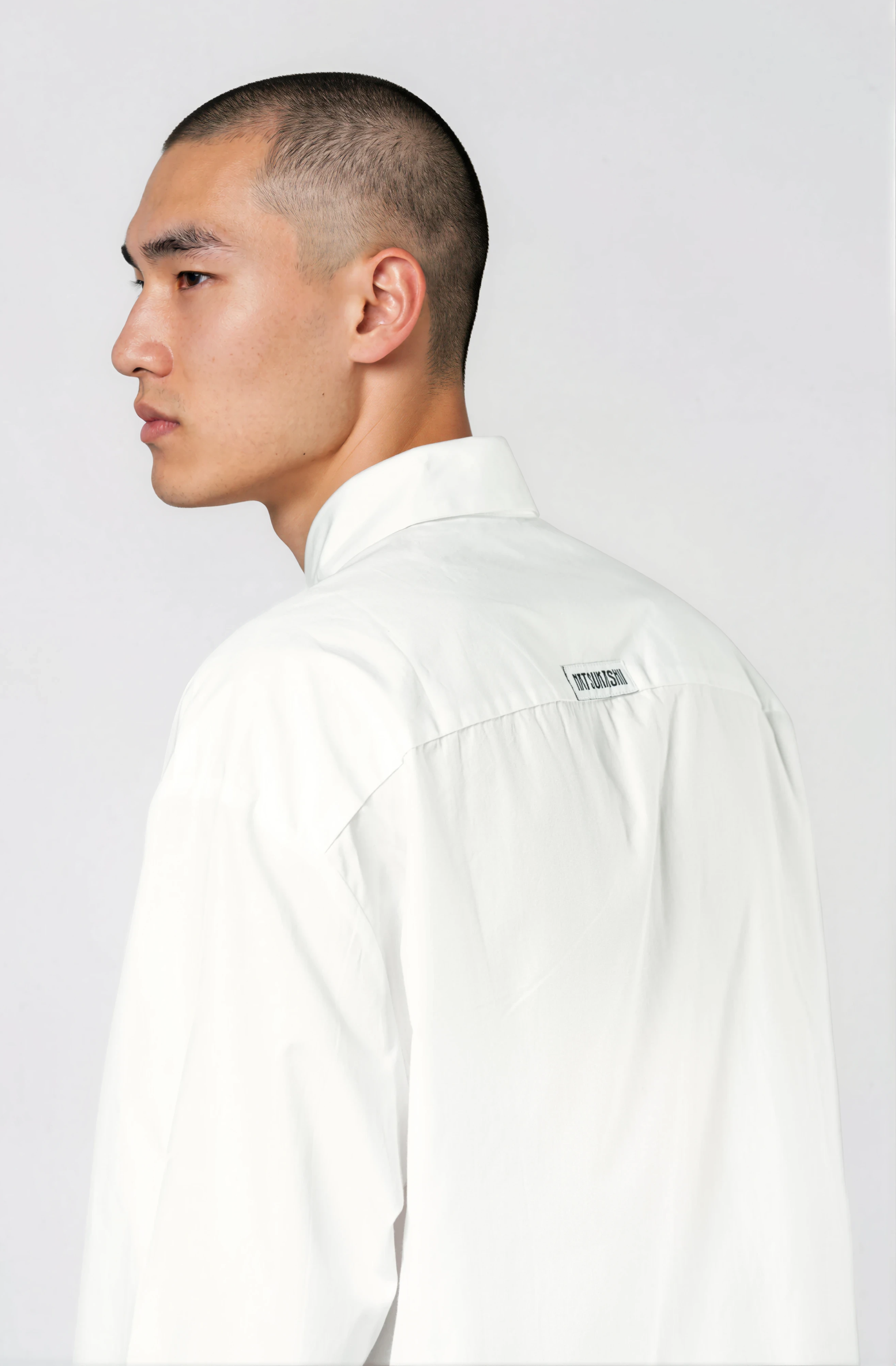A close-up profile of a man with a buzz cut hairstyle wearing a white shirt, standing against a light background.