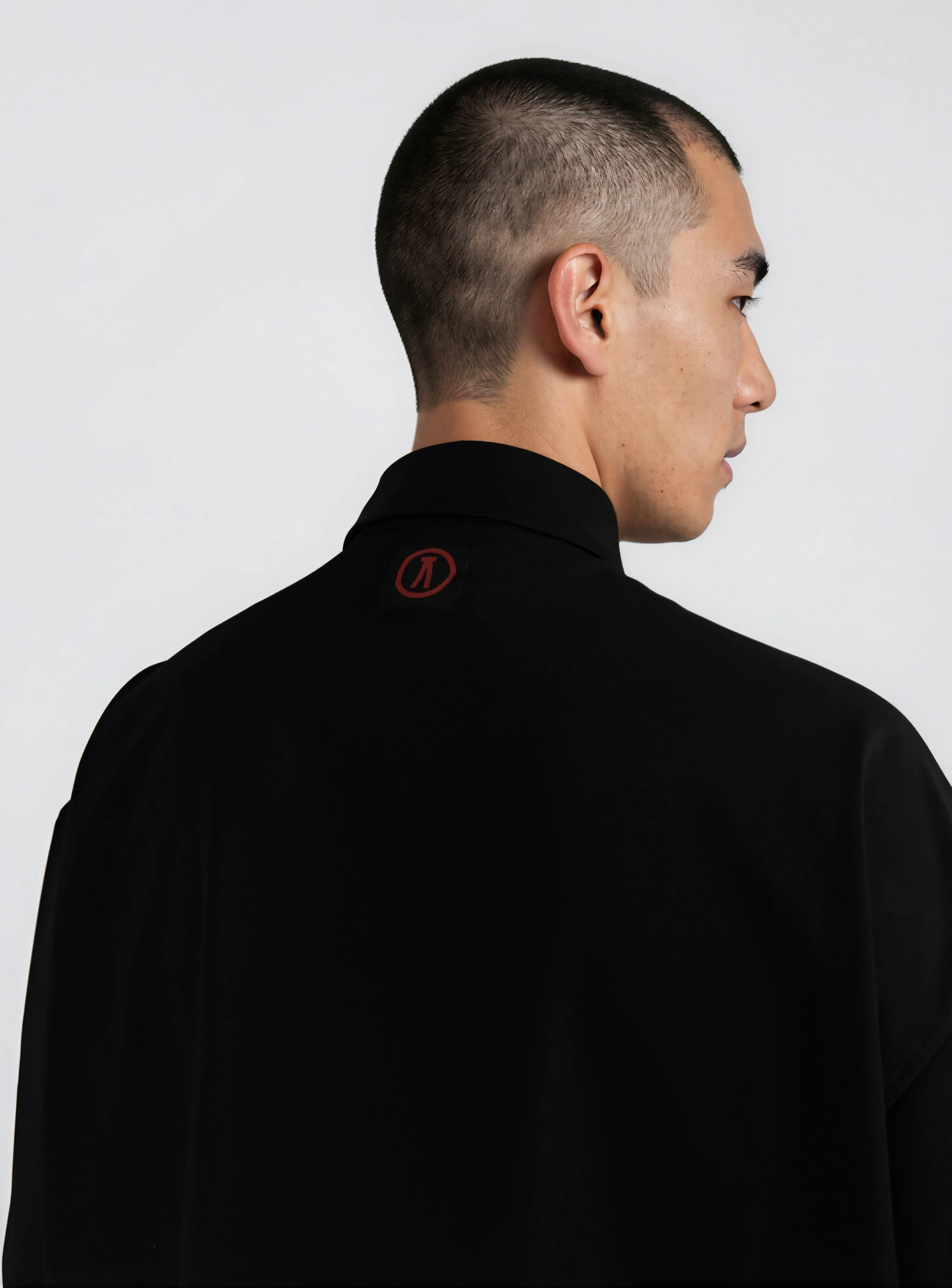 Side profile of a man with a short, buzzed haircut wearing a black shirt with a small red logo on the collar, standing against a plain white background.