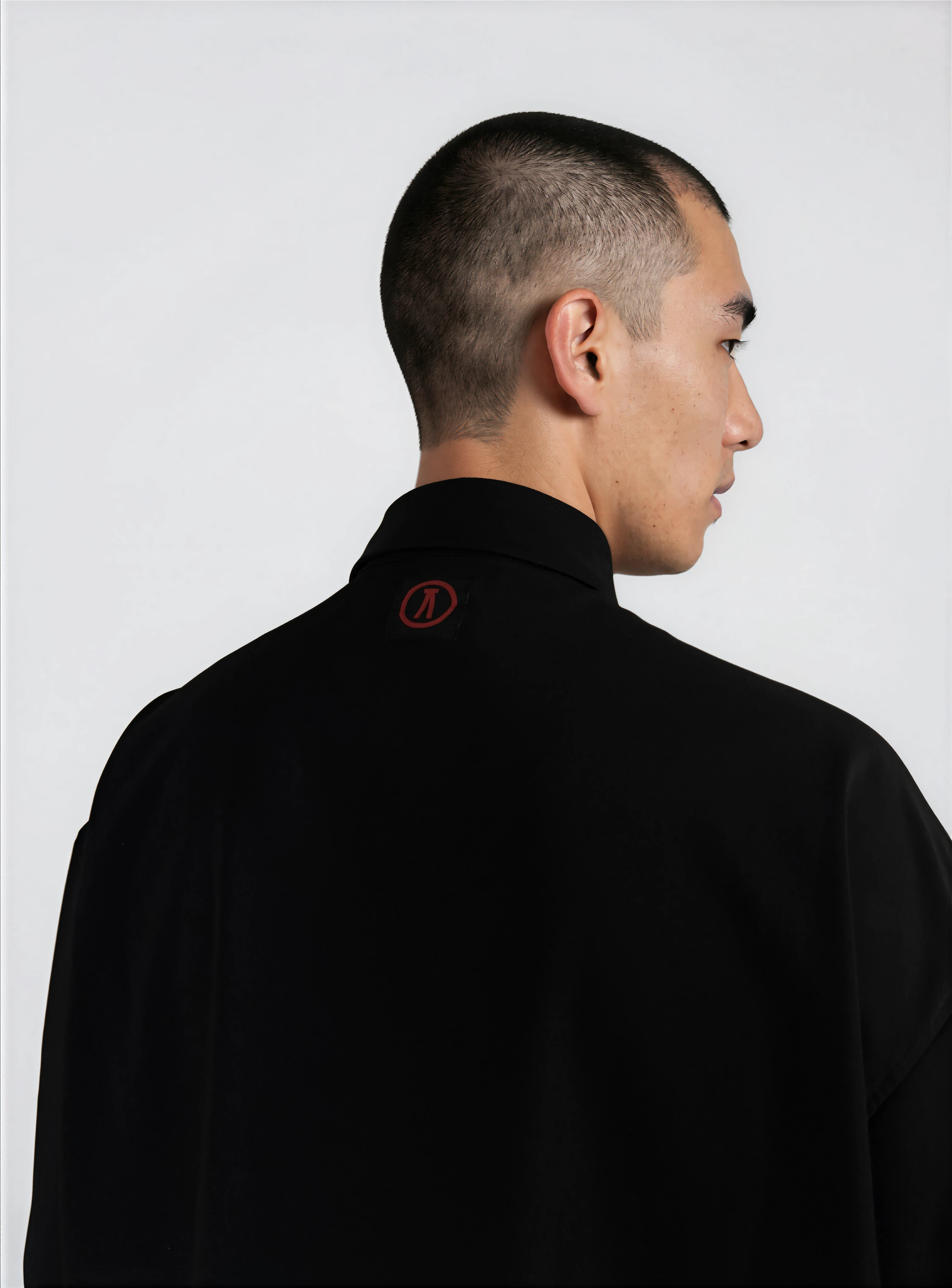Side profile of a man with a short, buzzed haircut wearing a black shirt with a small red logo on the collar, standing against a plain white background.
