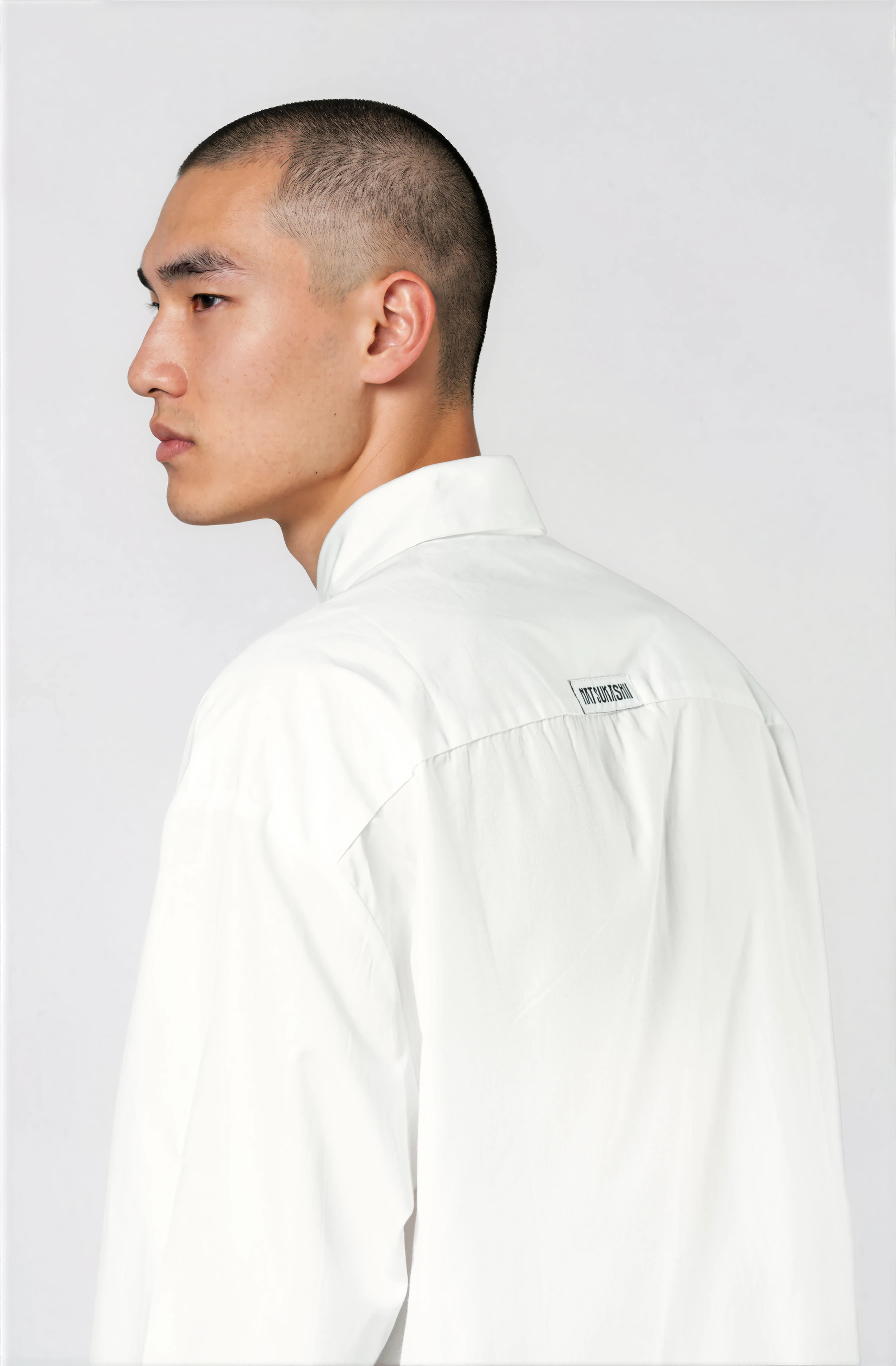 A close-up profile of a man with a buzz cut hairstyle wearing a white shirt, standing against a light background.