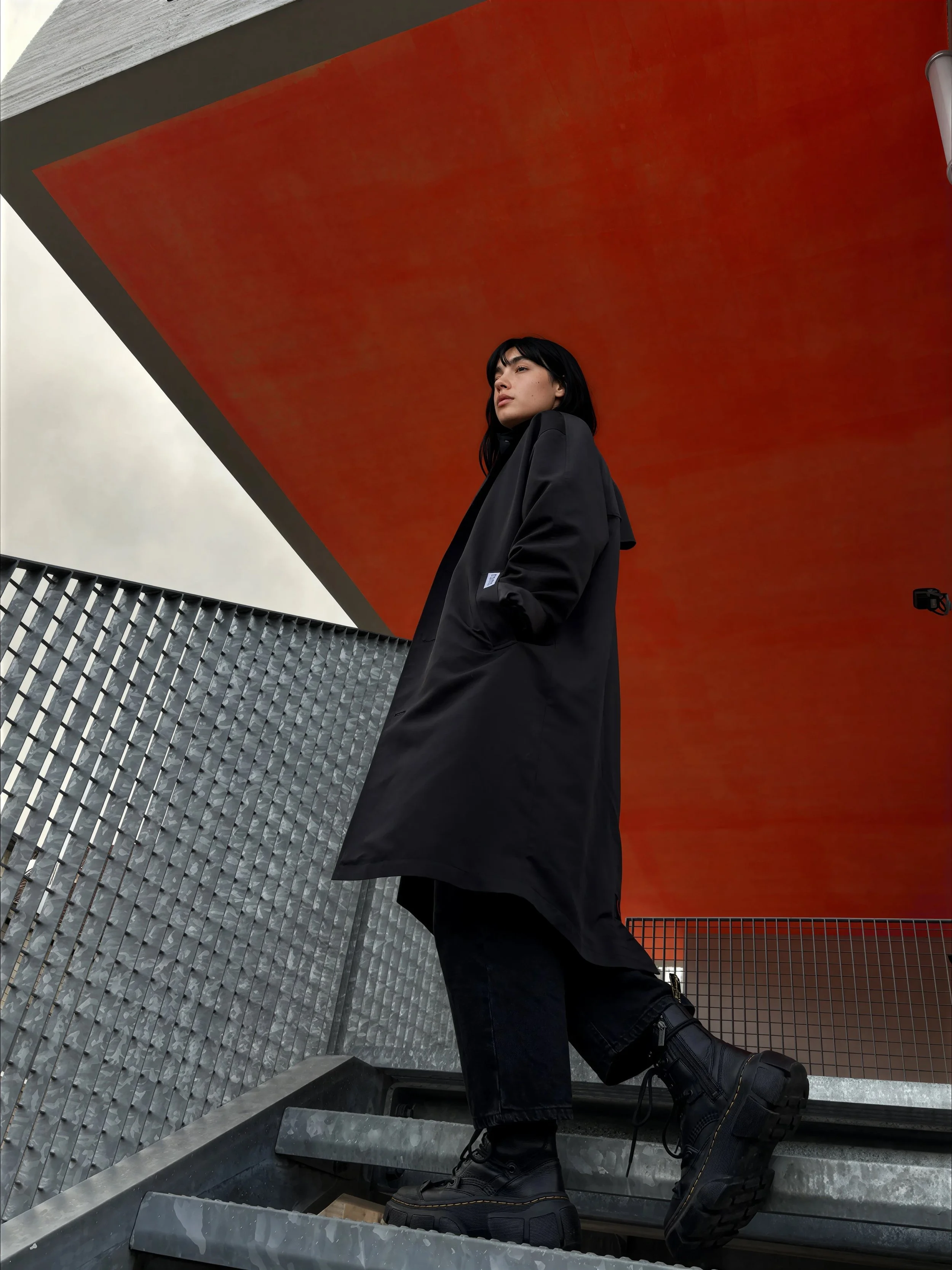 A woman with black hair, wearing a black jacket and black boots, kneeling on a staircase made of metal with textured side panels, under a large orange overhang with an overcast sky in the background.