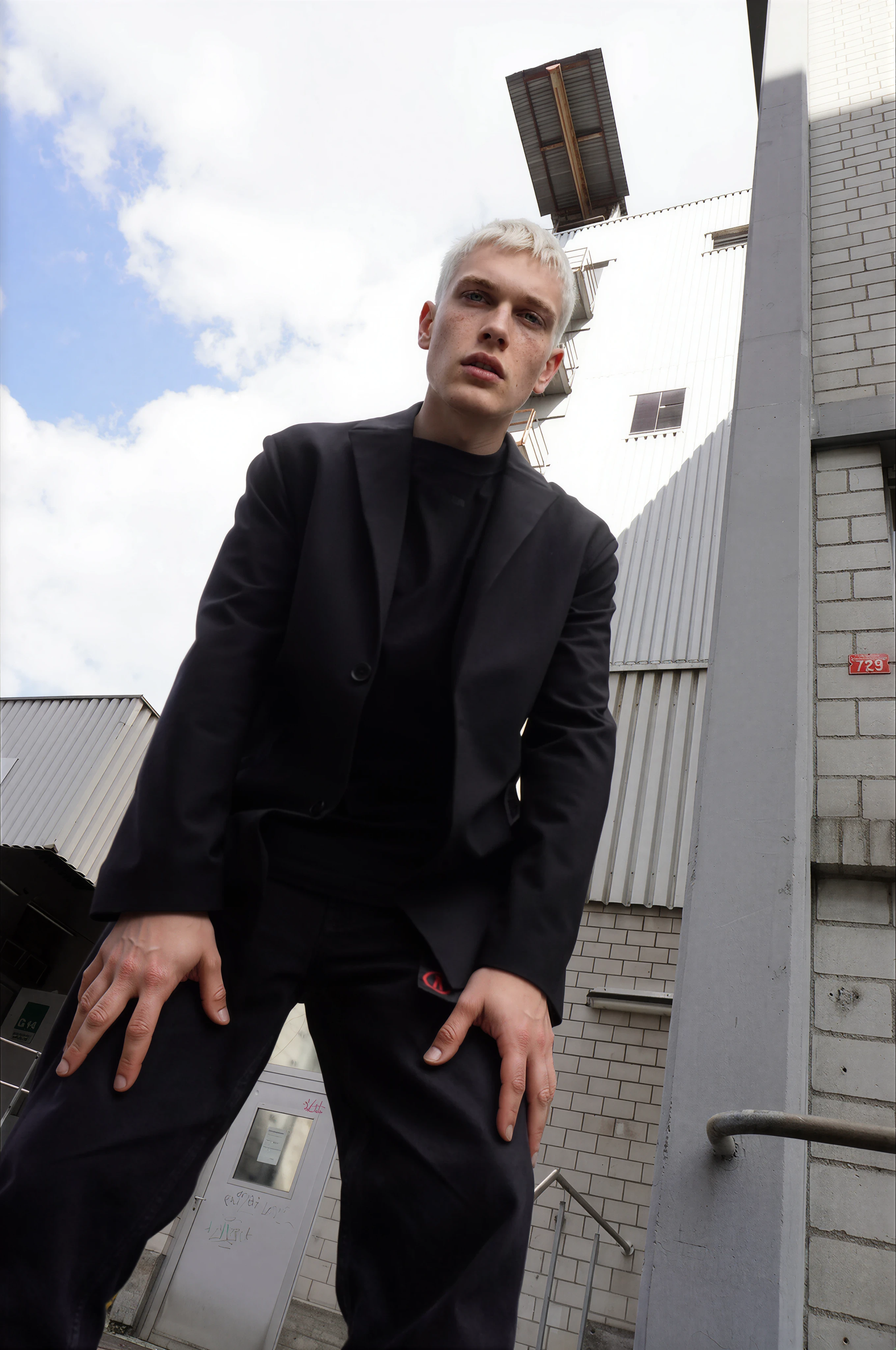 A young man with blond hair wearing a black blazer and black pants standing outdoors in front of a building with white brick and gray siding, looking at the camera with a serious expression, with clouds in the sky above.