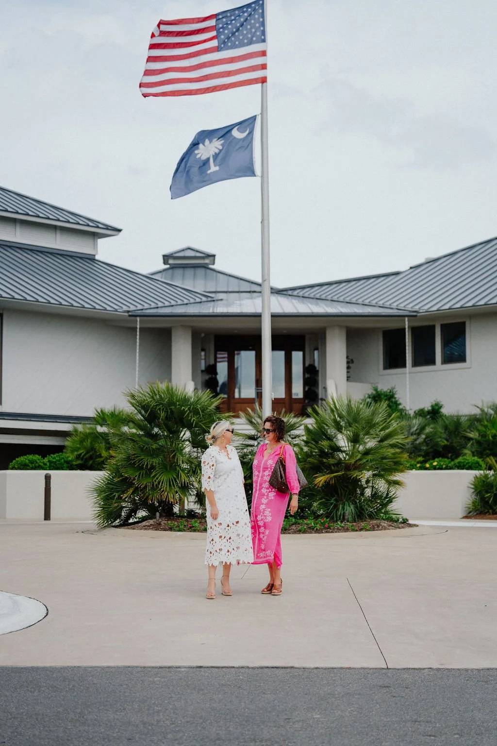 Two women stand and talk outside in front of a modern building with a flagpole flying the United States flag and a South Carolina state flag, surrounded by green bushes.