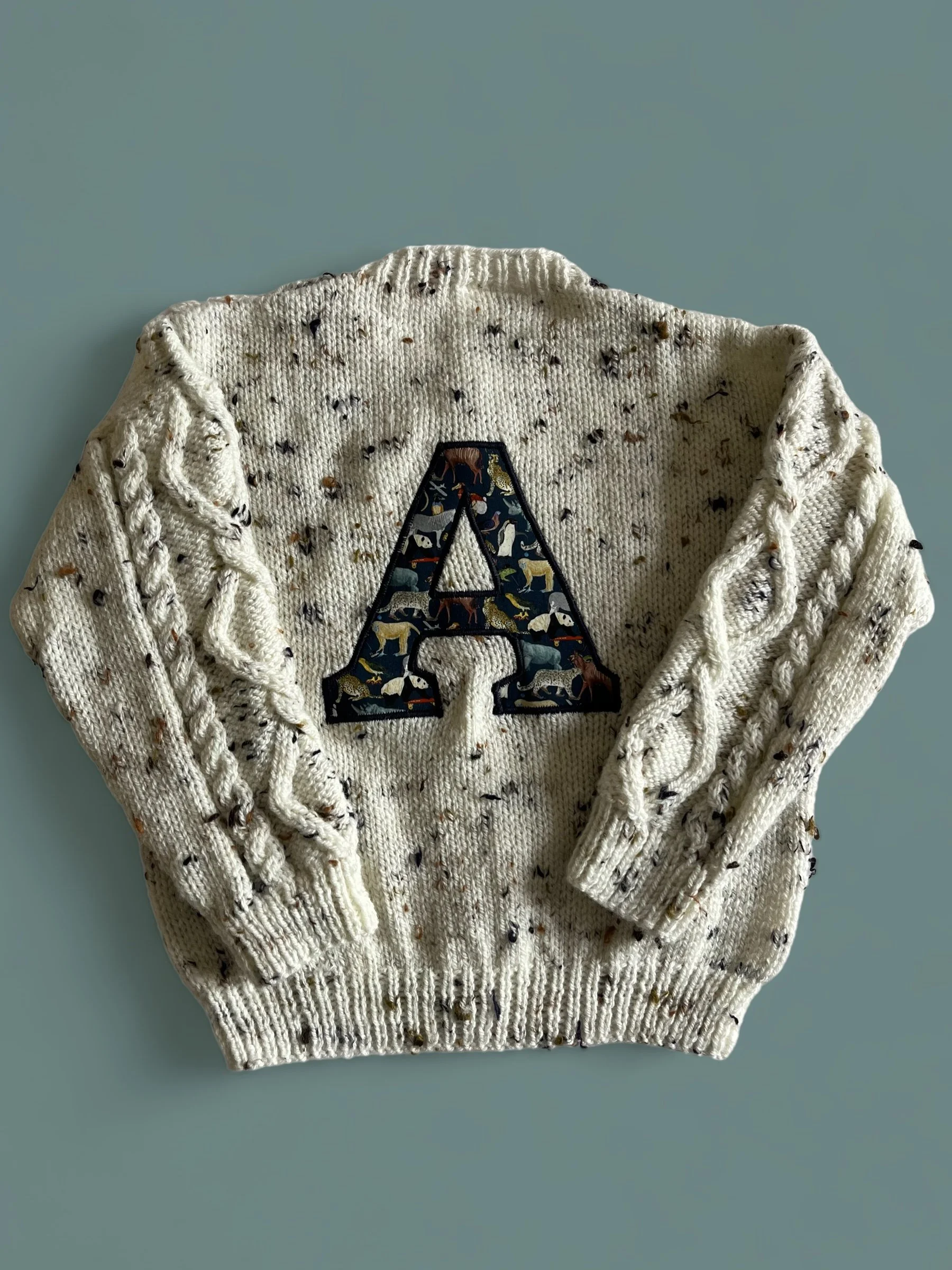 Cream-colored sweater with a large embroidered letter 'A' on the front, decorated with a pattern of various animals.