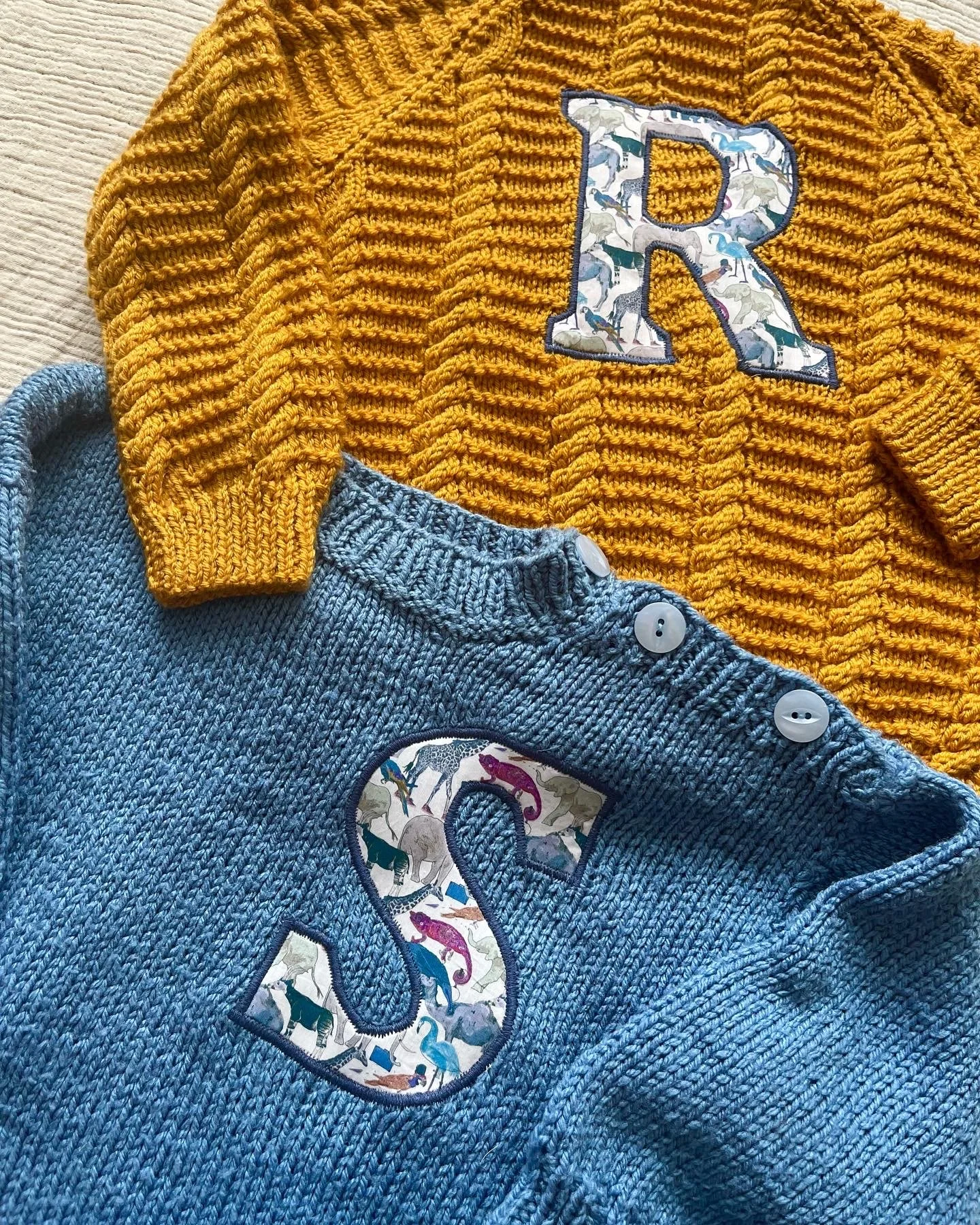 Two knitted children's sweaters with letter appliques, one mustard with 'R' and one blue with 'S' featuring animal patterns.