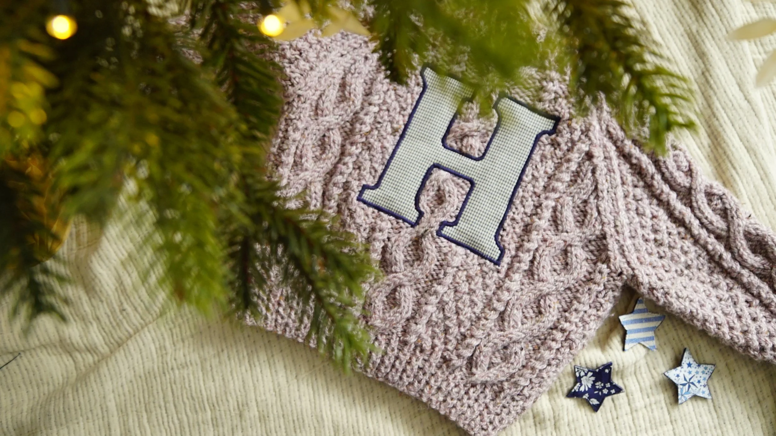 Gray knit sweater with a large letter 'H' embellishment, placed under evergreen branches and accompanied by decorative star shapes.