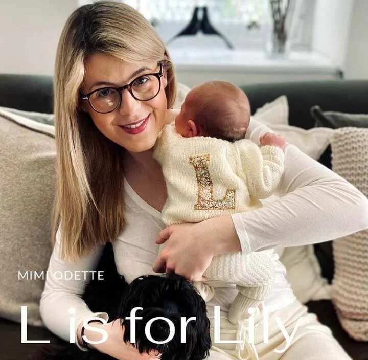 A woman with glasses holding a baby wearing a sweater featuring the letter 'L'. A black and white dog is also in her lap. Text on the image reads 'L is for Lily'. The setting appears to be a cozy indoor environment with cushions in the background.