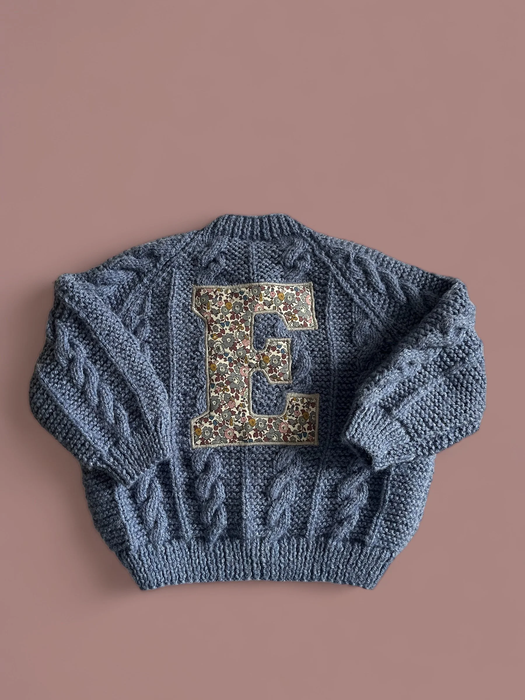 Gray knit sweater with cable patterns and a decorative floral letter E patch on the back.