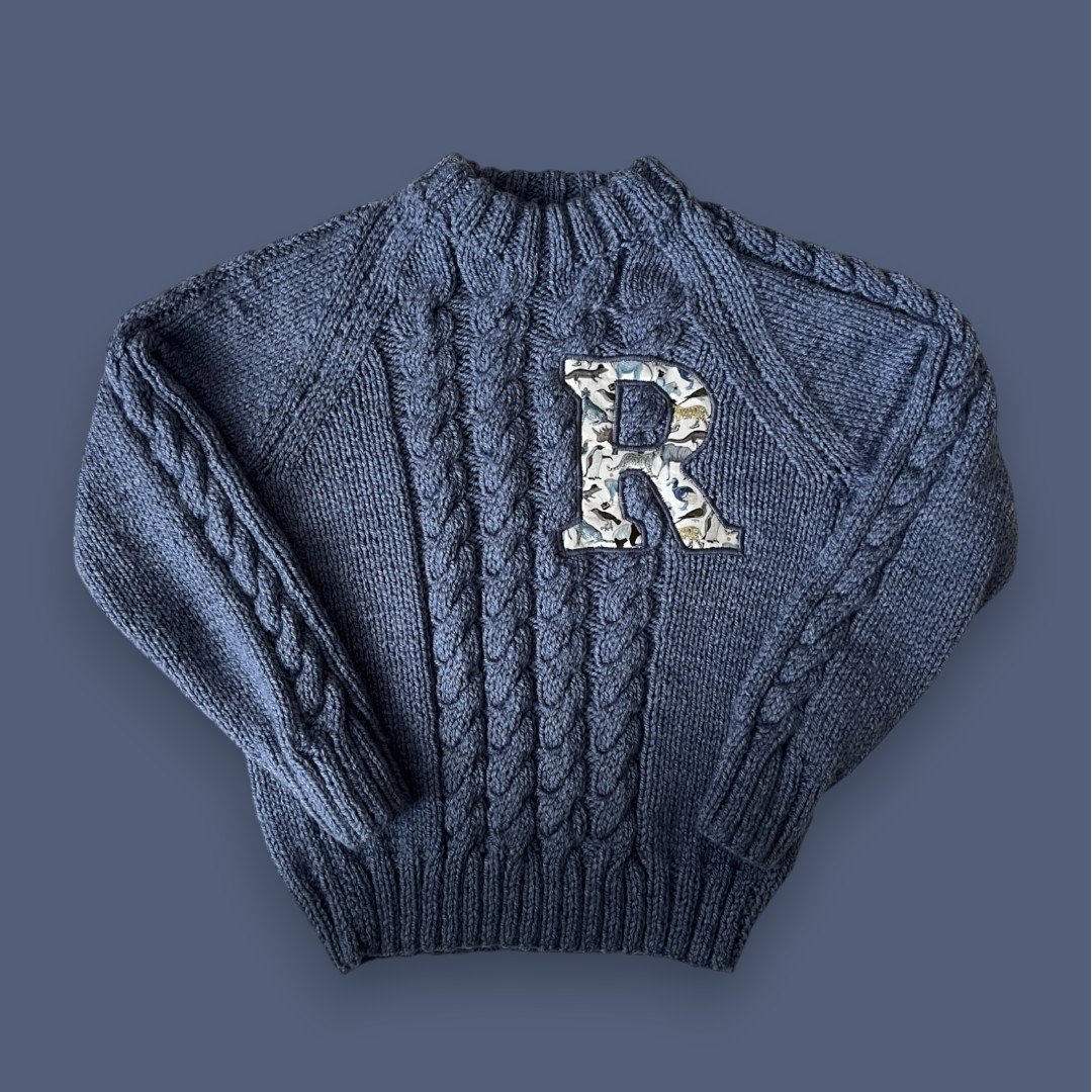 A gray knit sweater with cable patterns and an embroidered letter R on the front, displayed against a matching gray background.