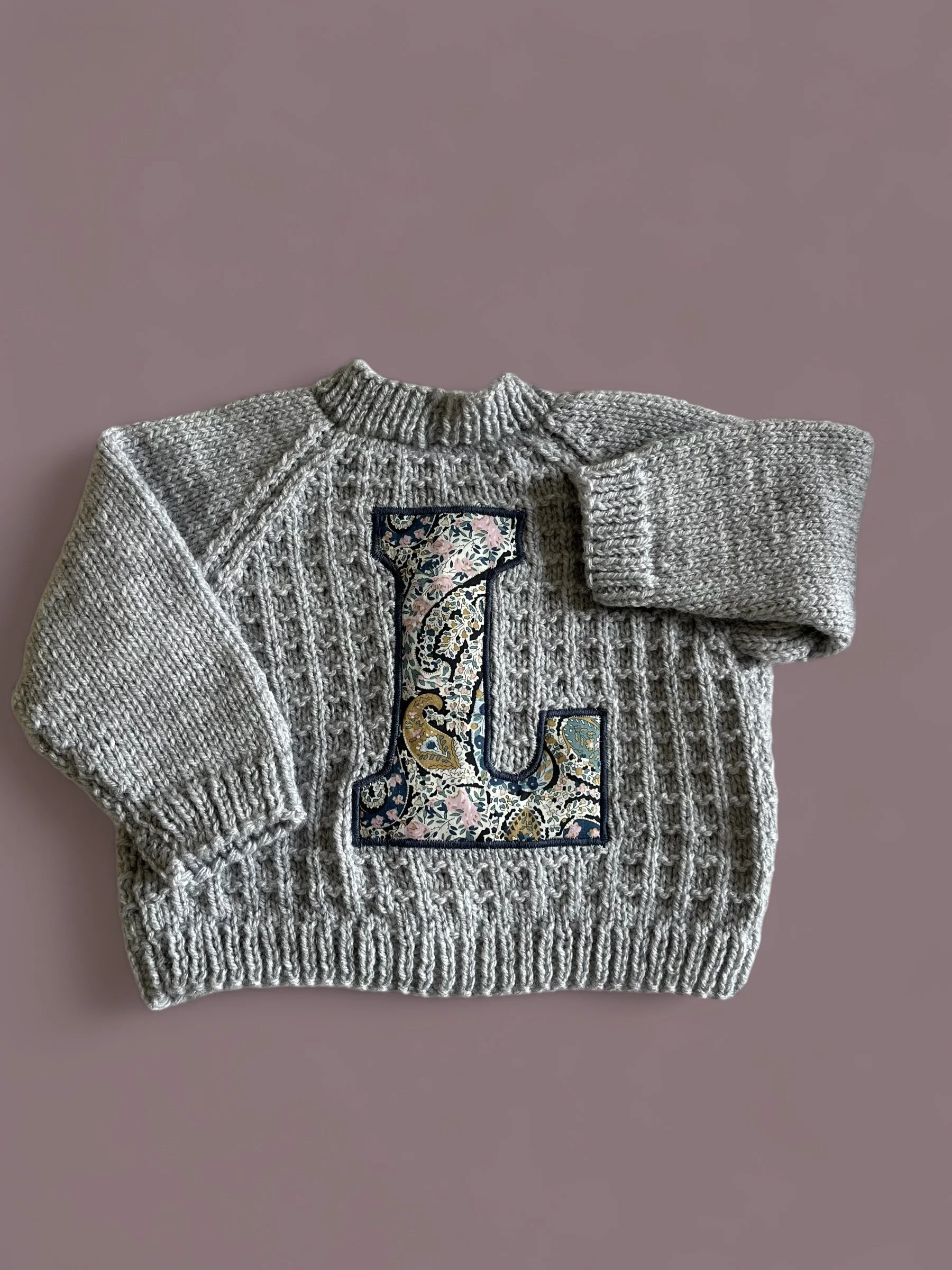 Gray knitted sweater with a floral patterned letter 'L' on the front, laid on a mauve surface.