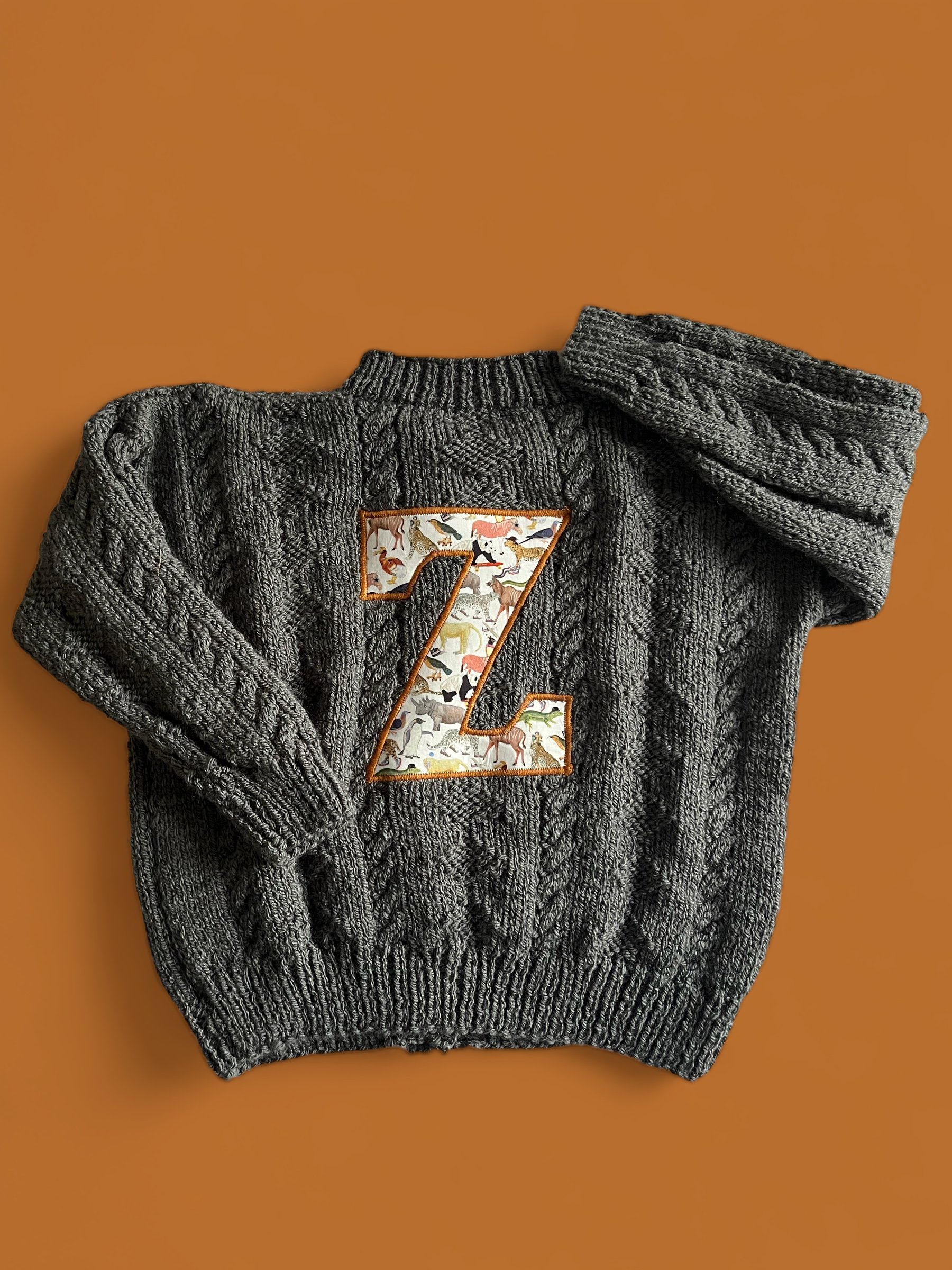 A gray knit sweater with a large letter Z patch featuring various small illustrations of animals.