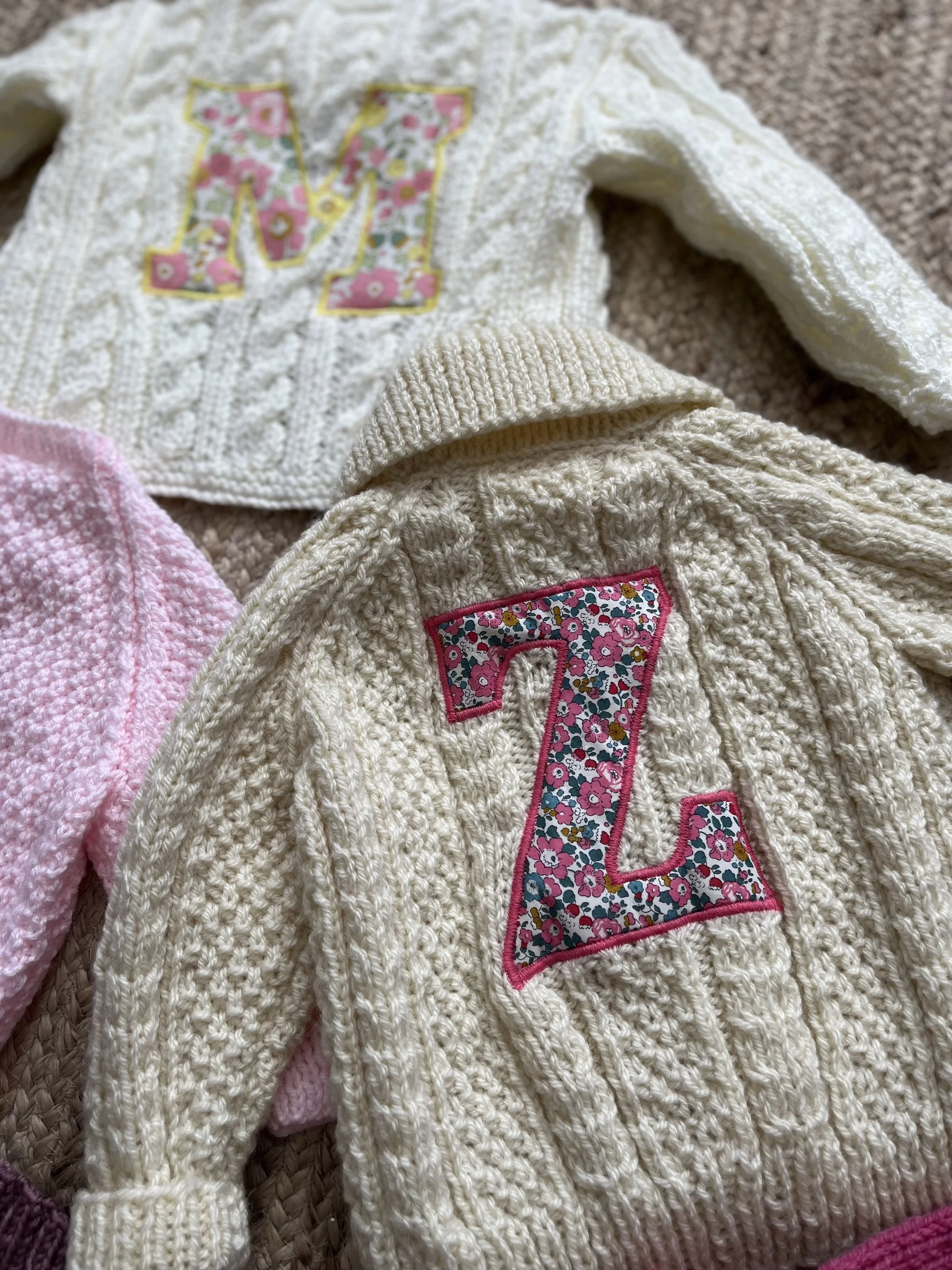 Three knitted sweaters, two cream-colored with floral letters 'M' and 'Z,' and one pink, arranged on a textile surface.