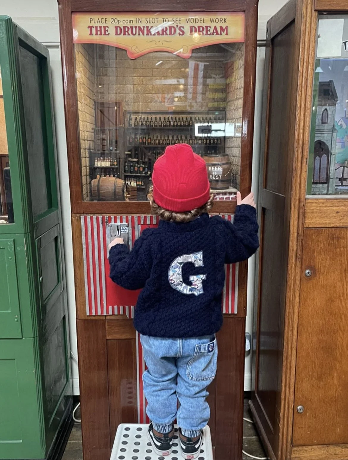 Child in a red hat and blue sweater with a large 'G' on the back, standing on a stool in front of a vintage arcade machine titled 'The Drunkard's Dream.'