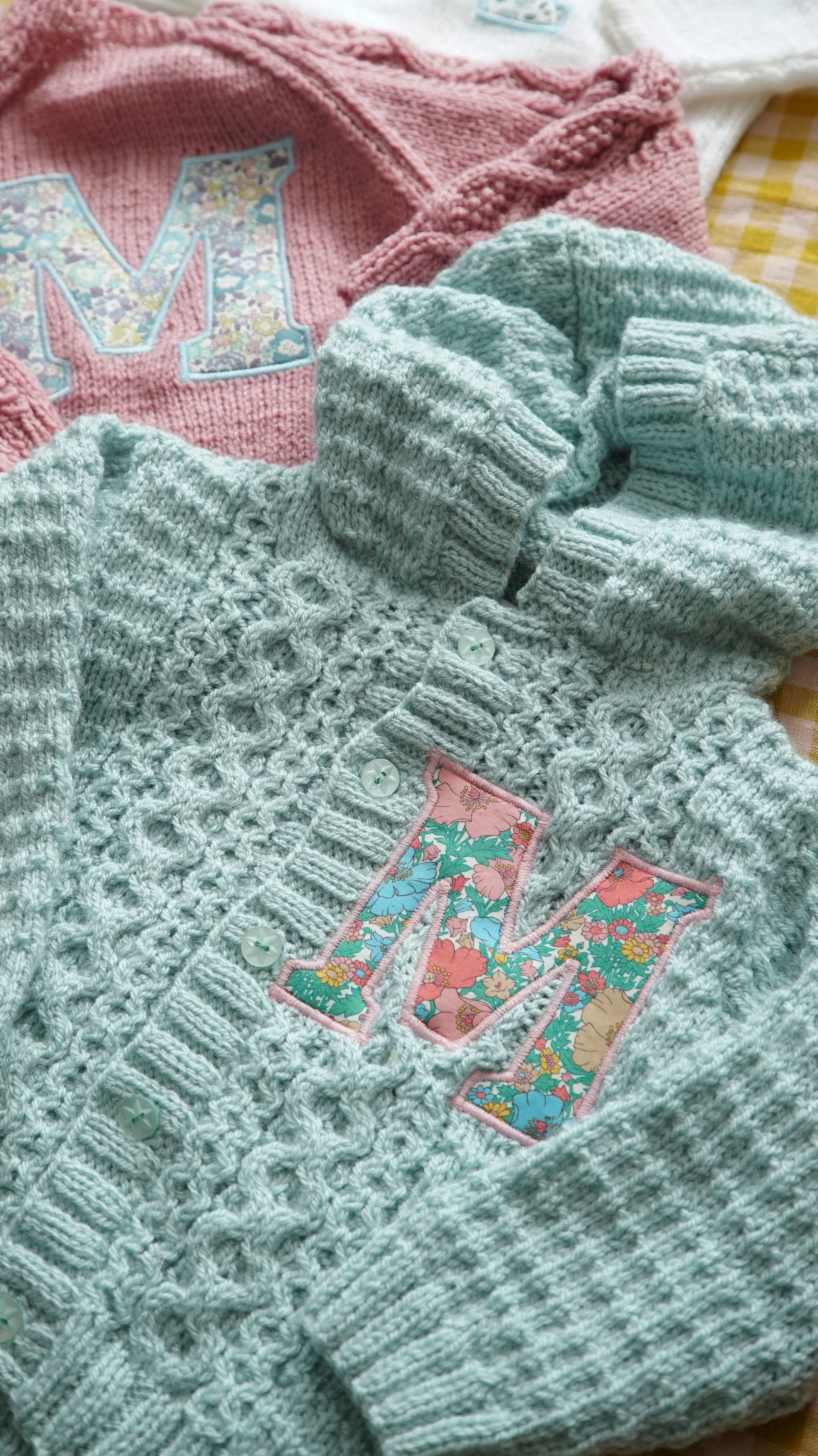Knitted children's cardigans with floral 'M' initials, one in mint green and one in pink.