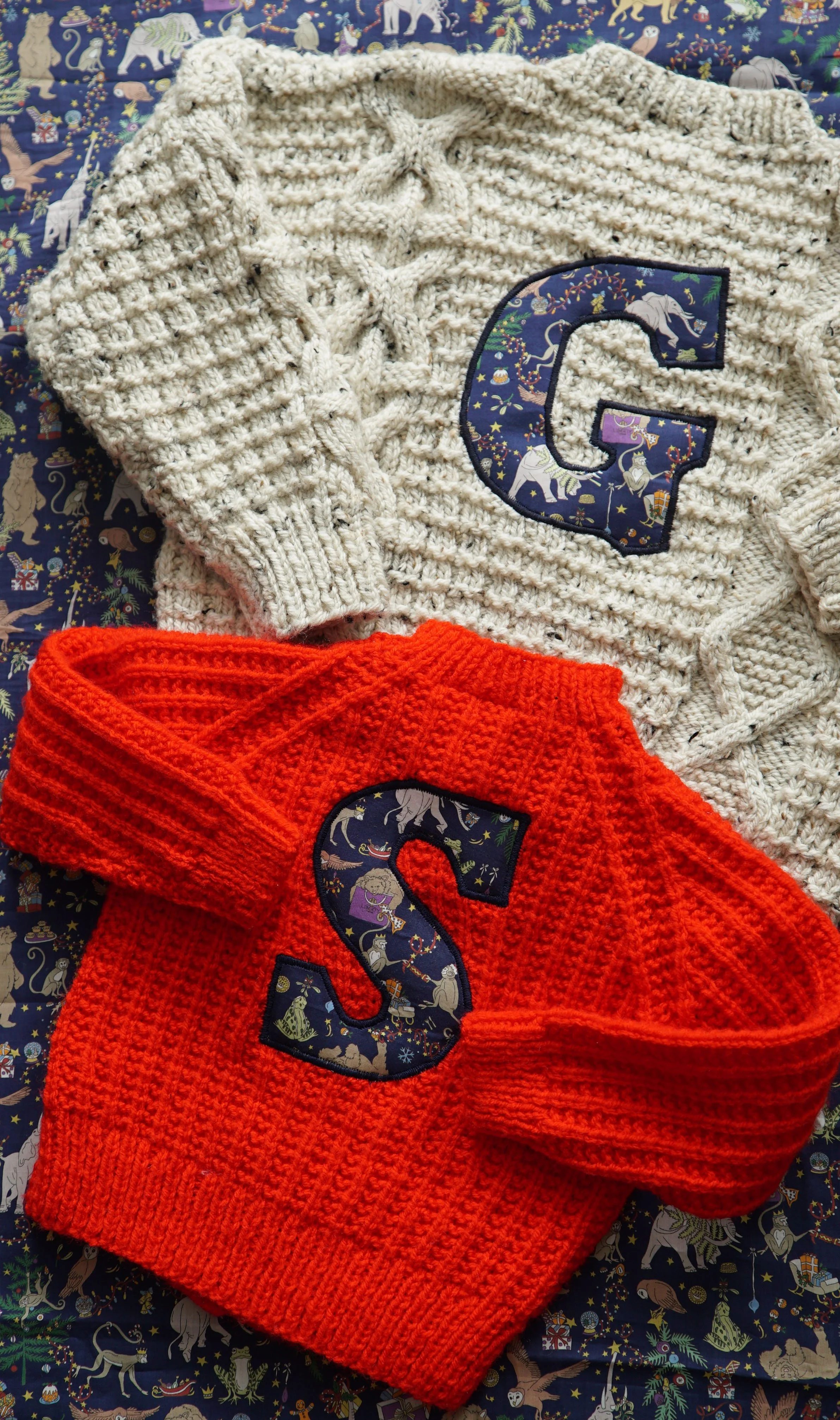 Two knitted sweaters with large letters are placed on a patterned fabric. One sweater is cream-colored with a 'G' and the other is red with an 'S'. Both letters feature a colorful animal and celestial print.