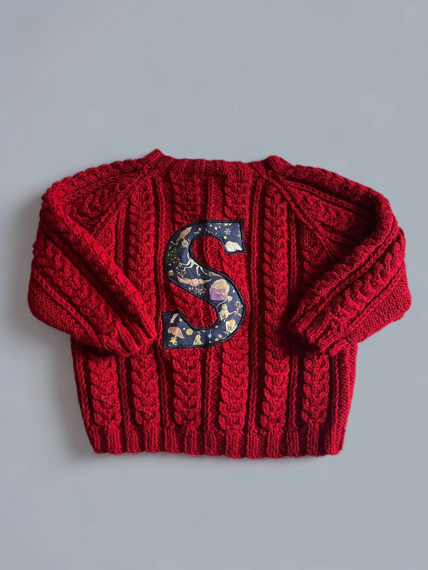 Red knitted sweater with a large embroidered letter 'S' on the front, decorated with celestial and animal motifs.