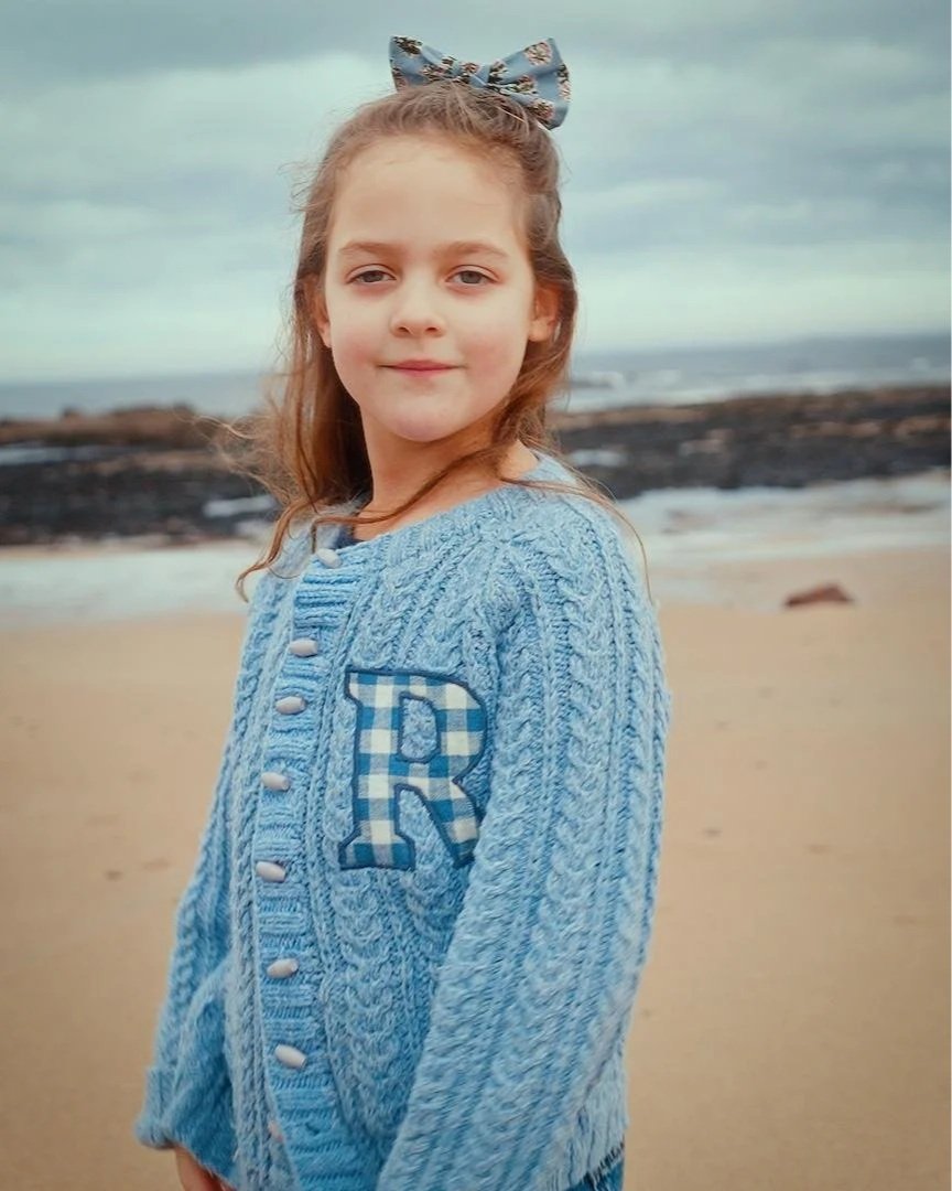Girl with a blue sweater and bow standing on a beach with cloudy sky.