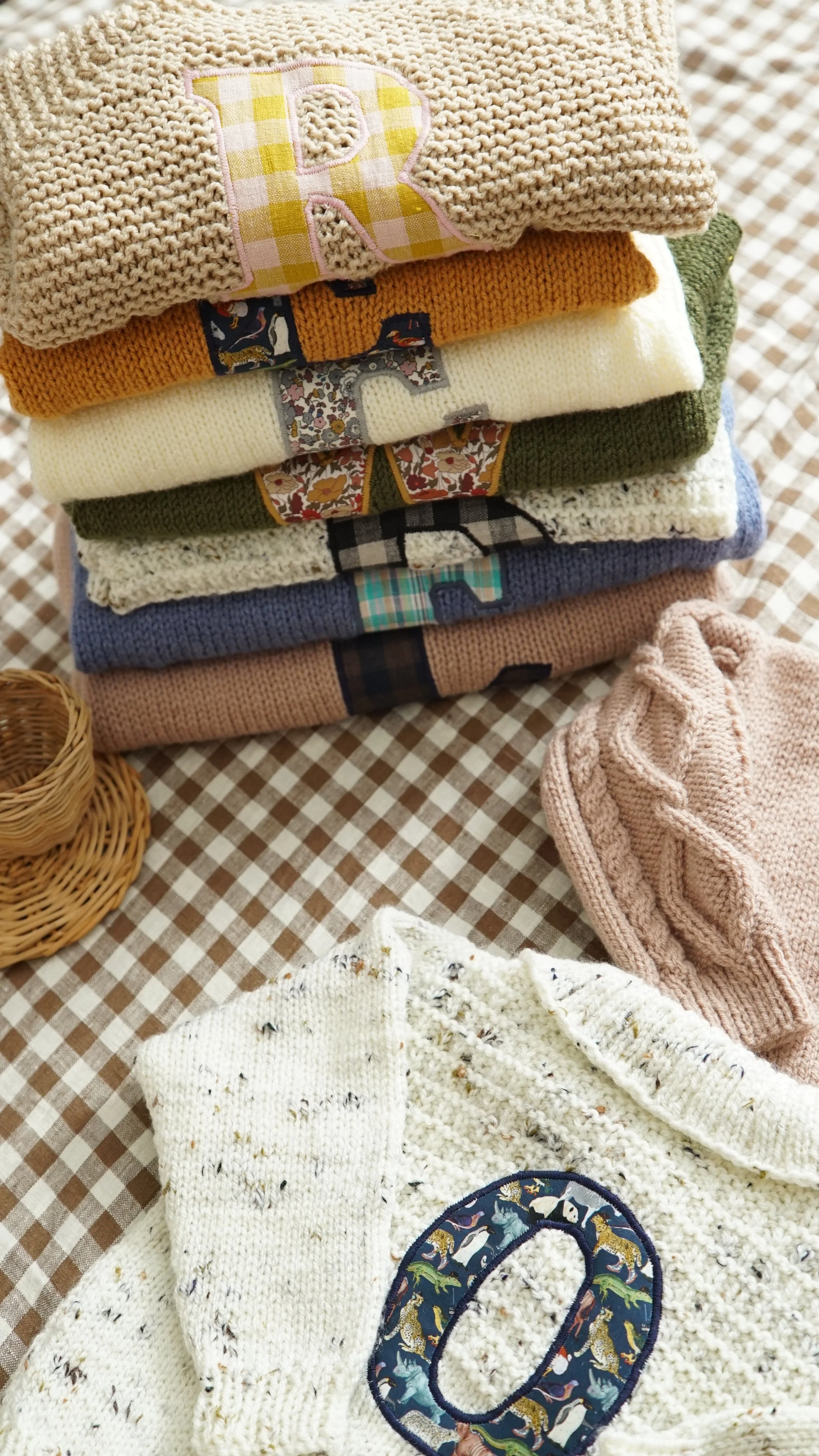 Stack of knitted sweaters with letters on them, folded on a checkered tablecloth, and a wicker basket beside them.